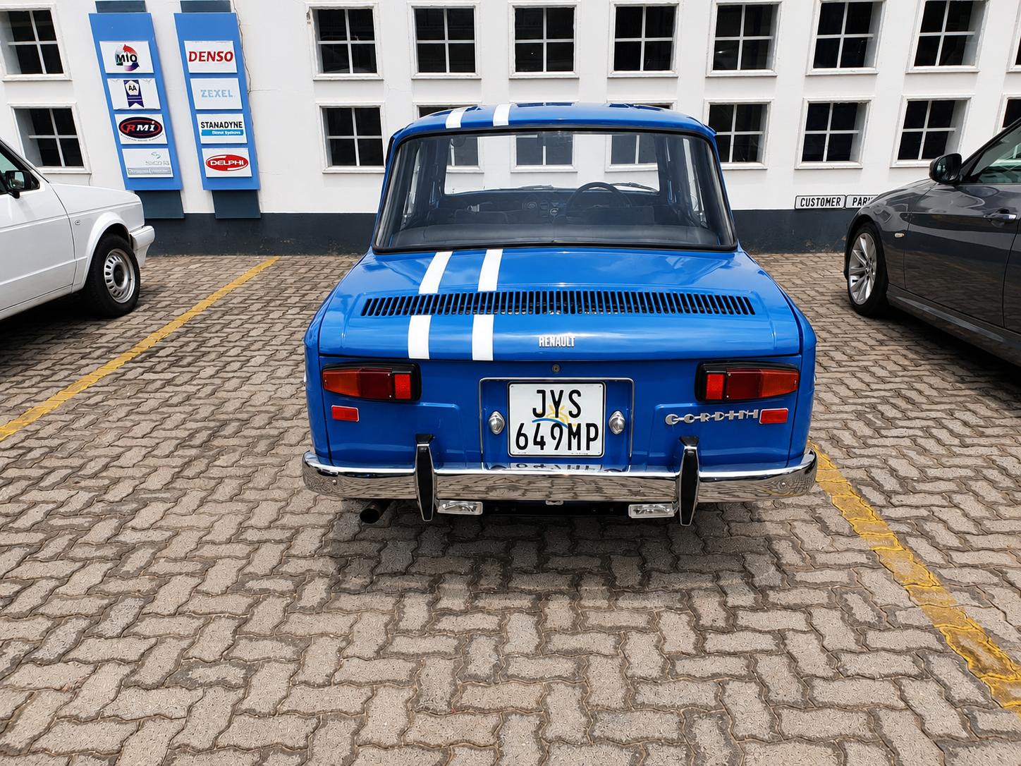 A vintage blue Renault GORDINI sports car with white racing stripes parked in a parking lot, with a white building in the background and two other cars on each side.