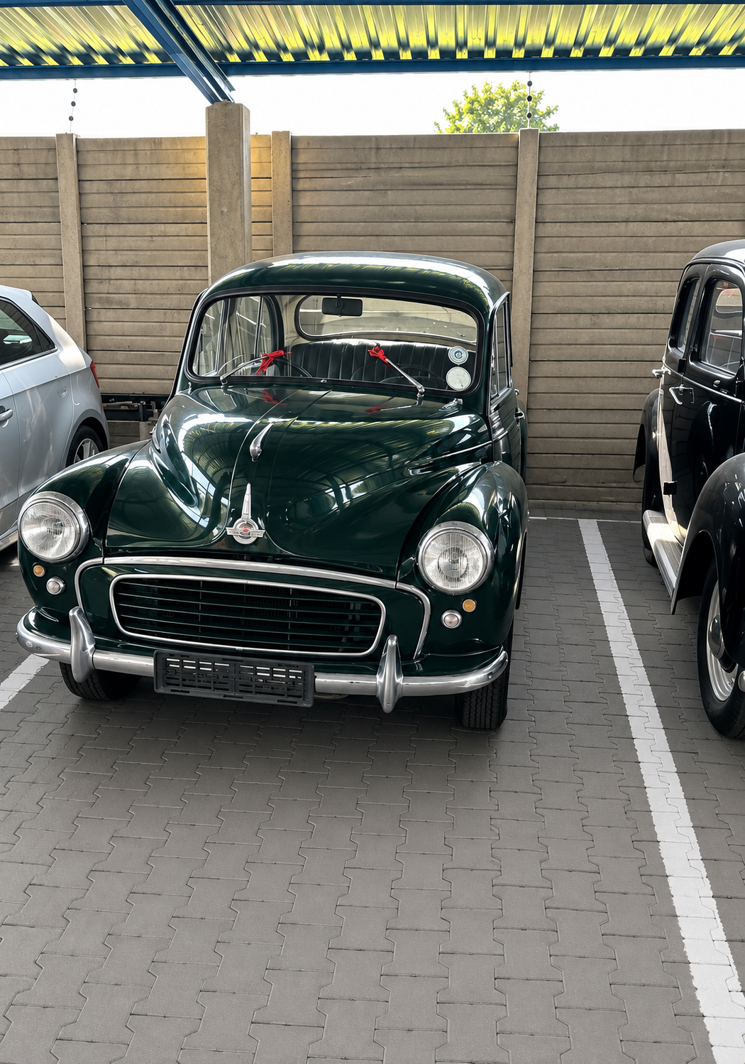 A vintage dark green car with a rounded body and large headlights parked in an indoor parking space, with a beige wooden fence and partial view of other cars around.