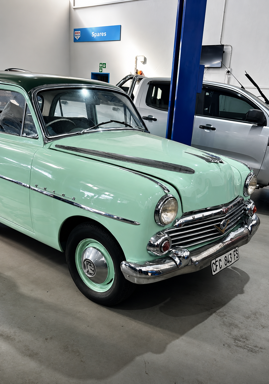 A vintage mint green Ford car parked inside a garage with a blue sign that reads 'Spares' and a silver pickup truck in the background.