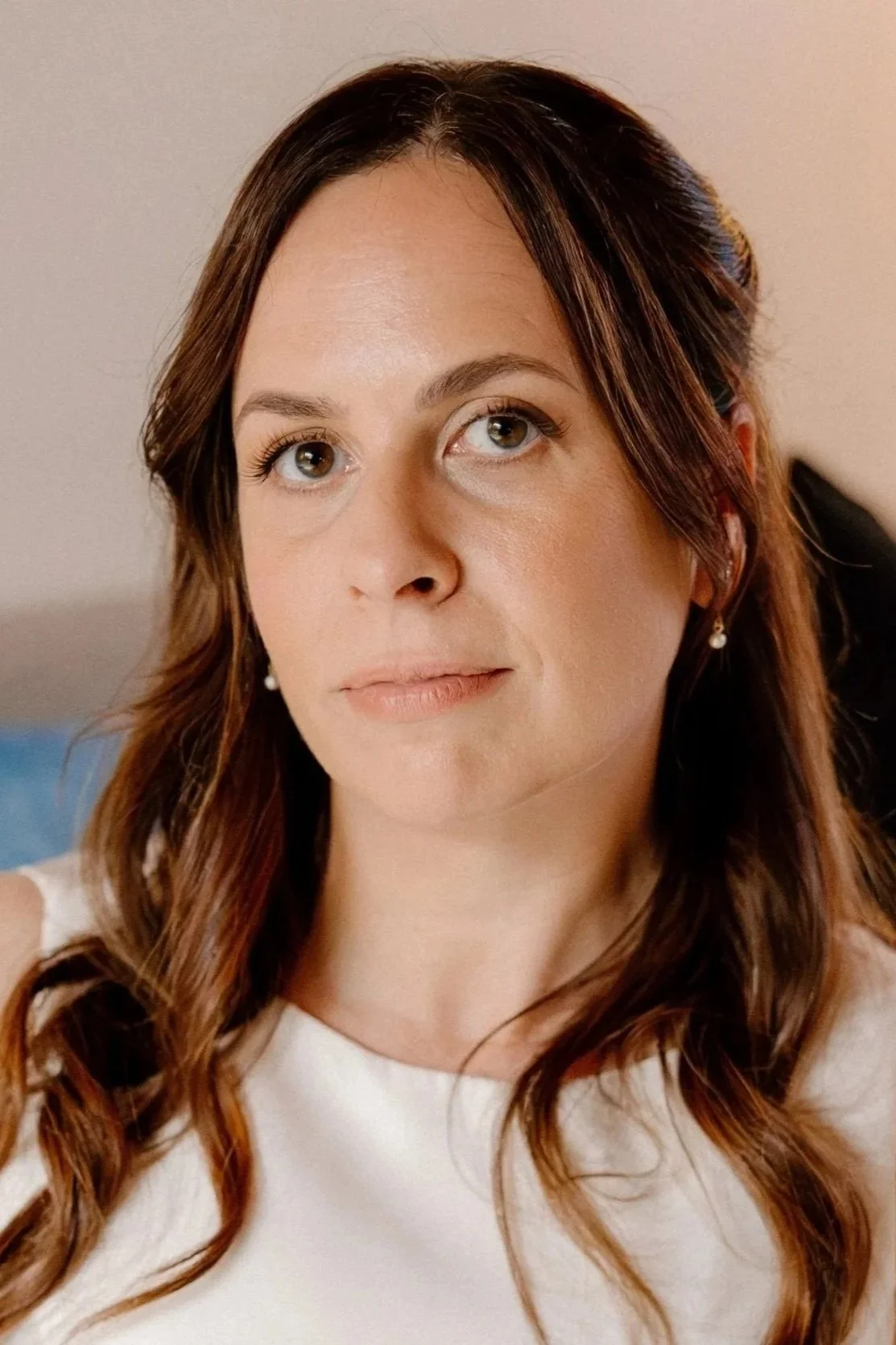 A woman with brown hair styled in loose curls, wearing a white top, small pearl earrings, looking slightly to her left with a neutral expression.