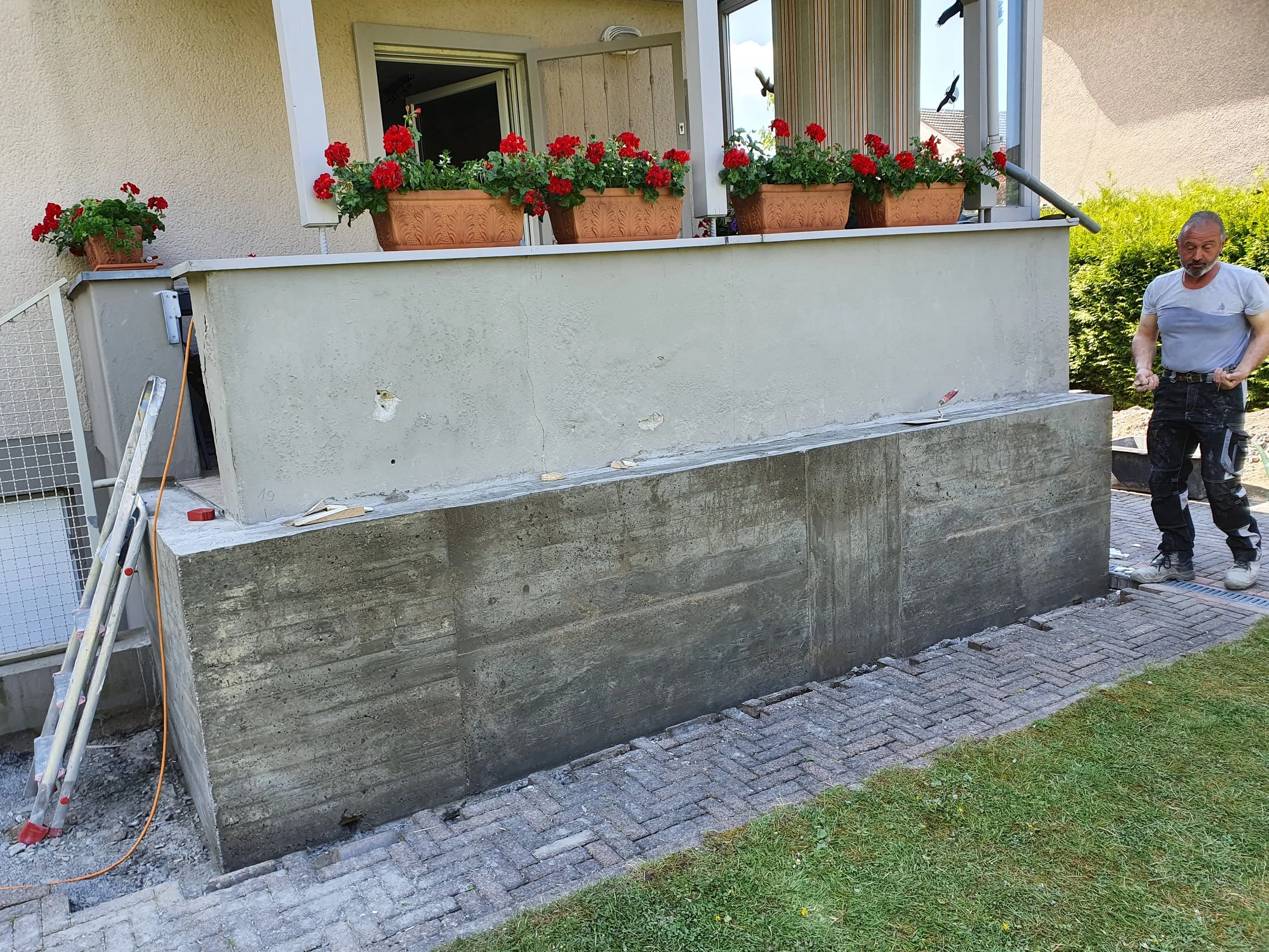 Ein Mann steht neben einer unvollendeten Hausverkleidung auf einer Terrasse, umgeben von Bauutensilien. Vor ihm befindet sich eine Mauer, die noch bearbeitet wird, mit blauen Abdeckungen im oberen Bereich. An der Wand sind Blumentöpfe mit roten Blumen angebracht.