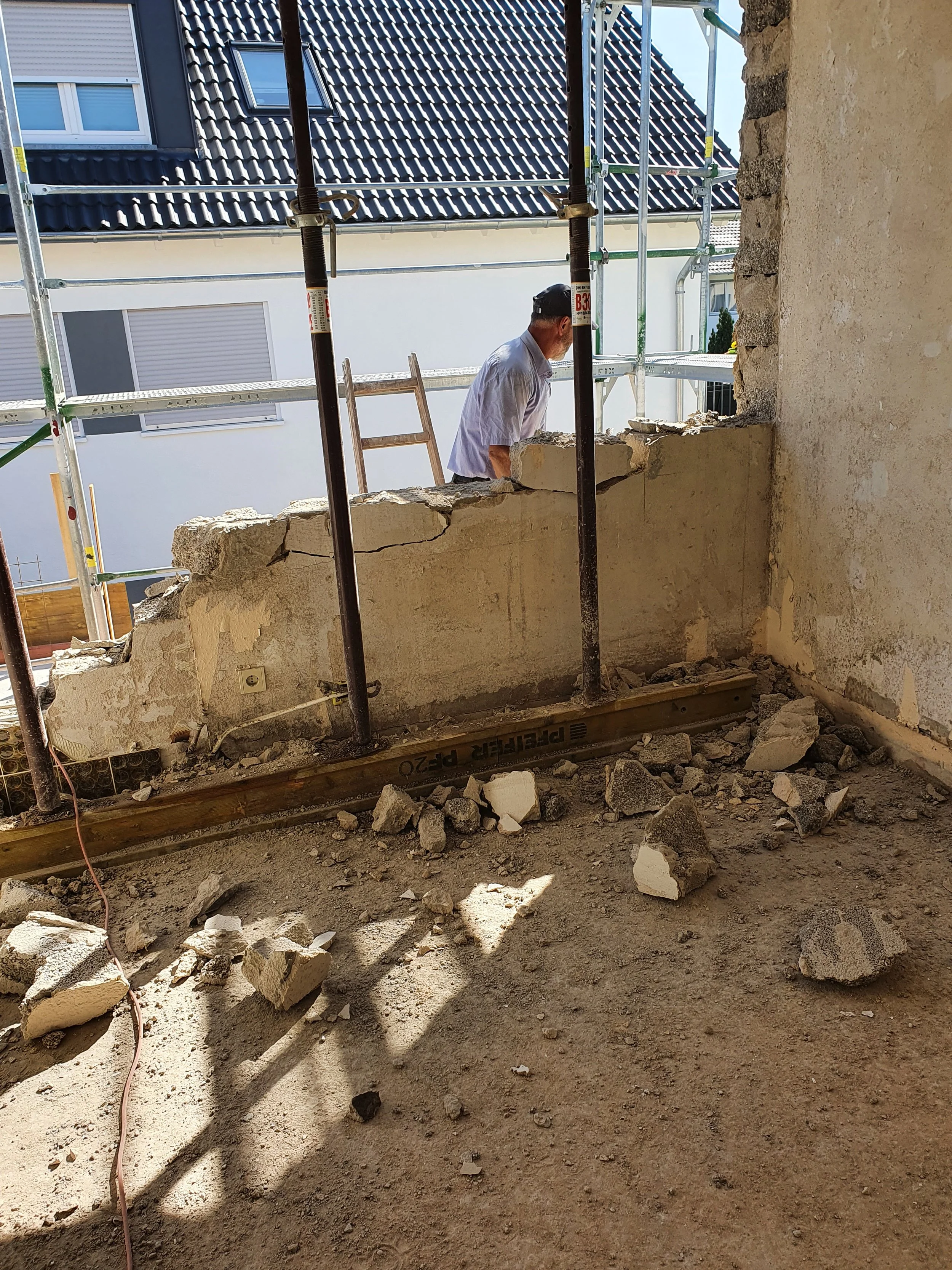 Ein Mann arbeitet an einer Baustelle, bei der eine Wand abgerissen wird. Es gibt Gerüste im Hintergrund und Trümmer auf dem Boden.