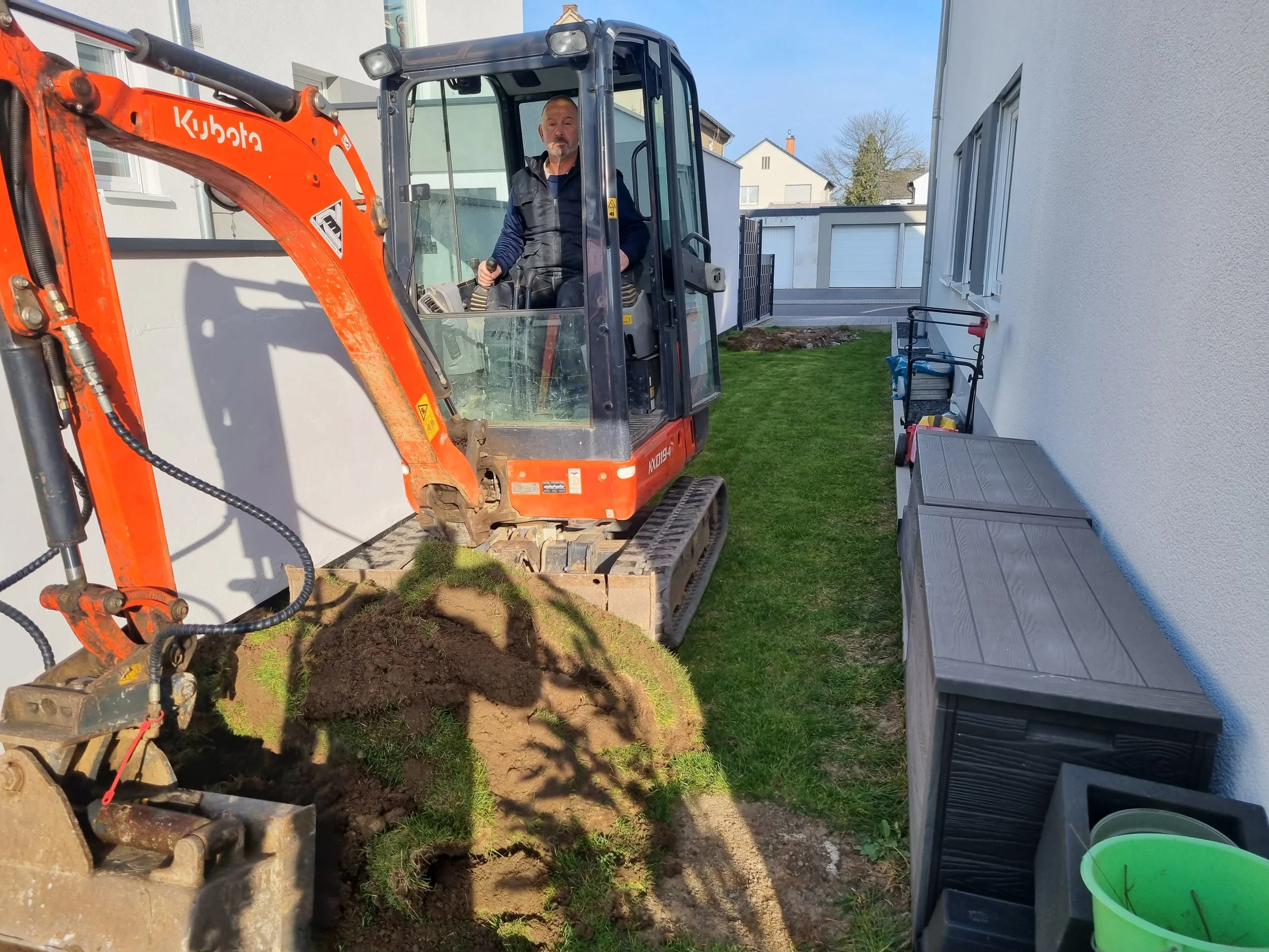 Mann bedient Mini-Bagger beim Graben neben einem weißen Haus, mit Gartenwerkzeugen und Gartenmöbeln im Hintergrund.