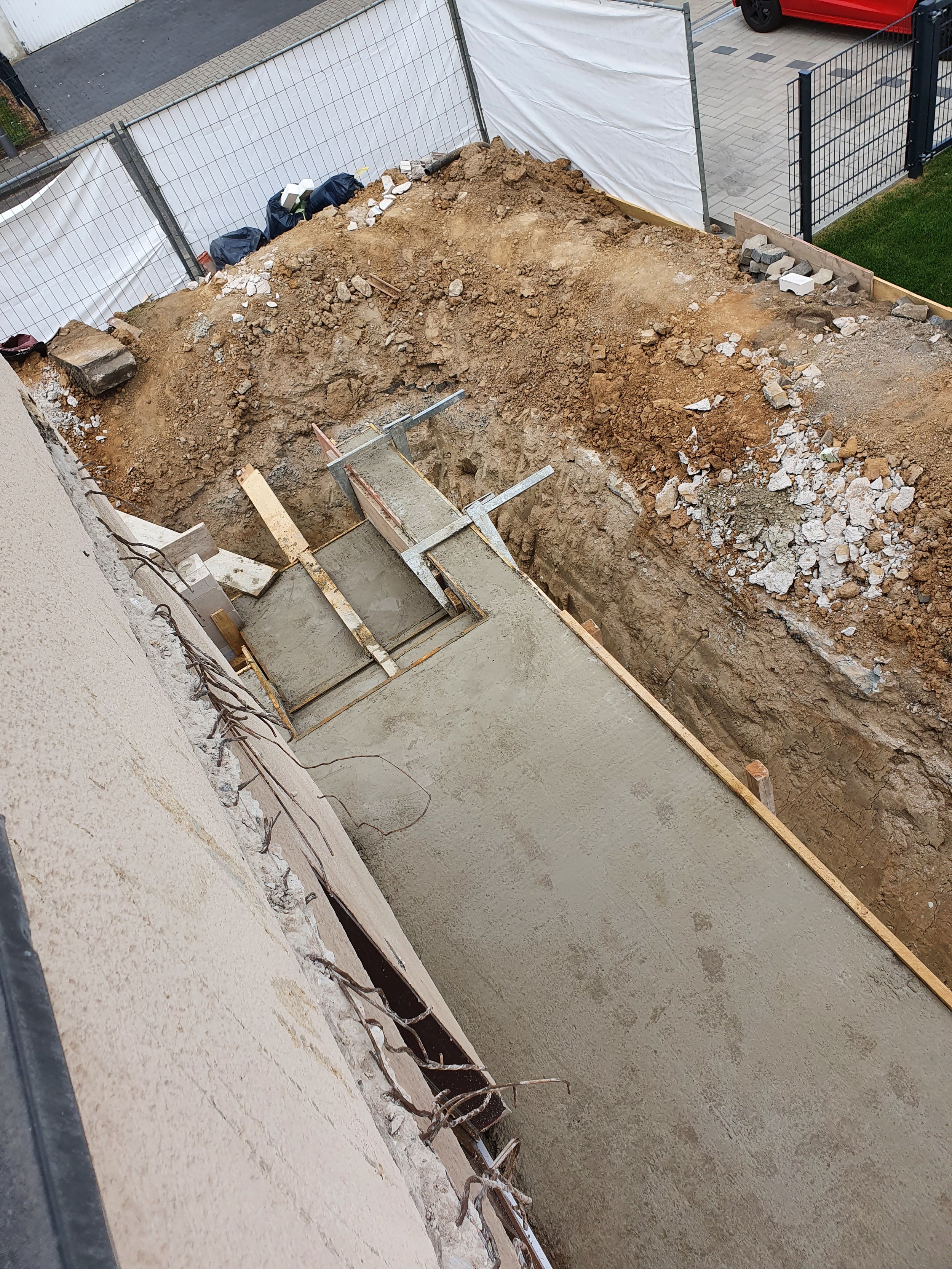 Baugrube vor einem Haus mit frischer Betonfläche, umgeben von Baugerüsten und Zäunen.