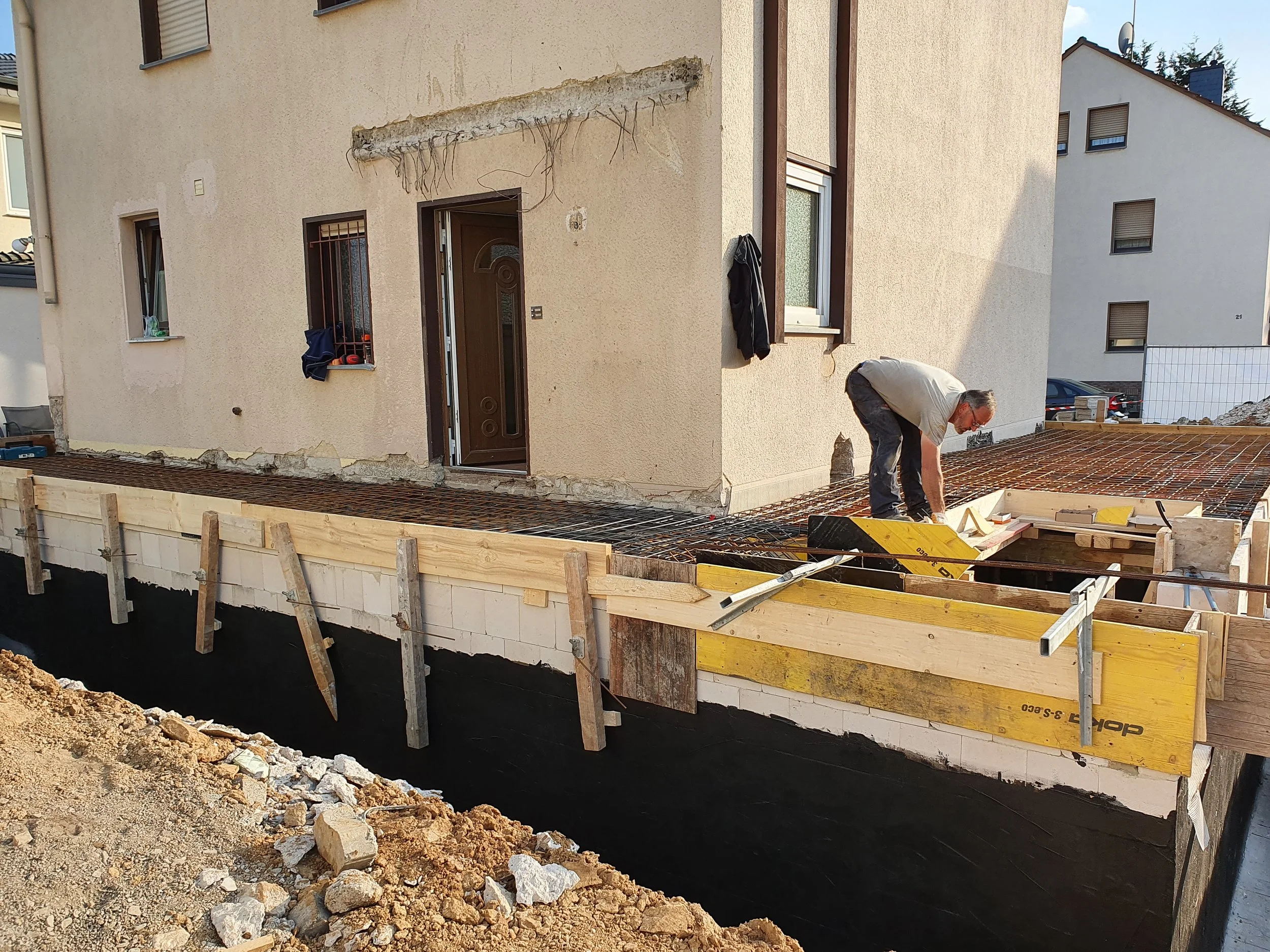 Ein Bauarbeiter arbeitet auf einer Terrasse vor einem Mehrfamilienhaus. Die Terrasse wird gerade errichtet, mit Stahlbewehrung und Holzrahmen.