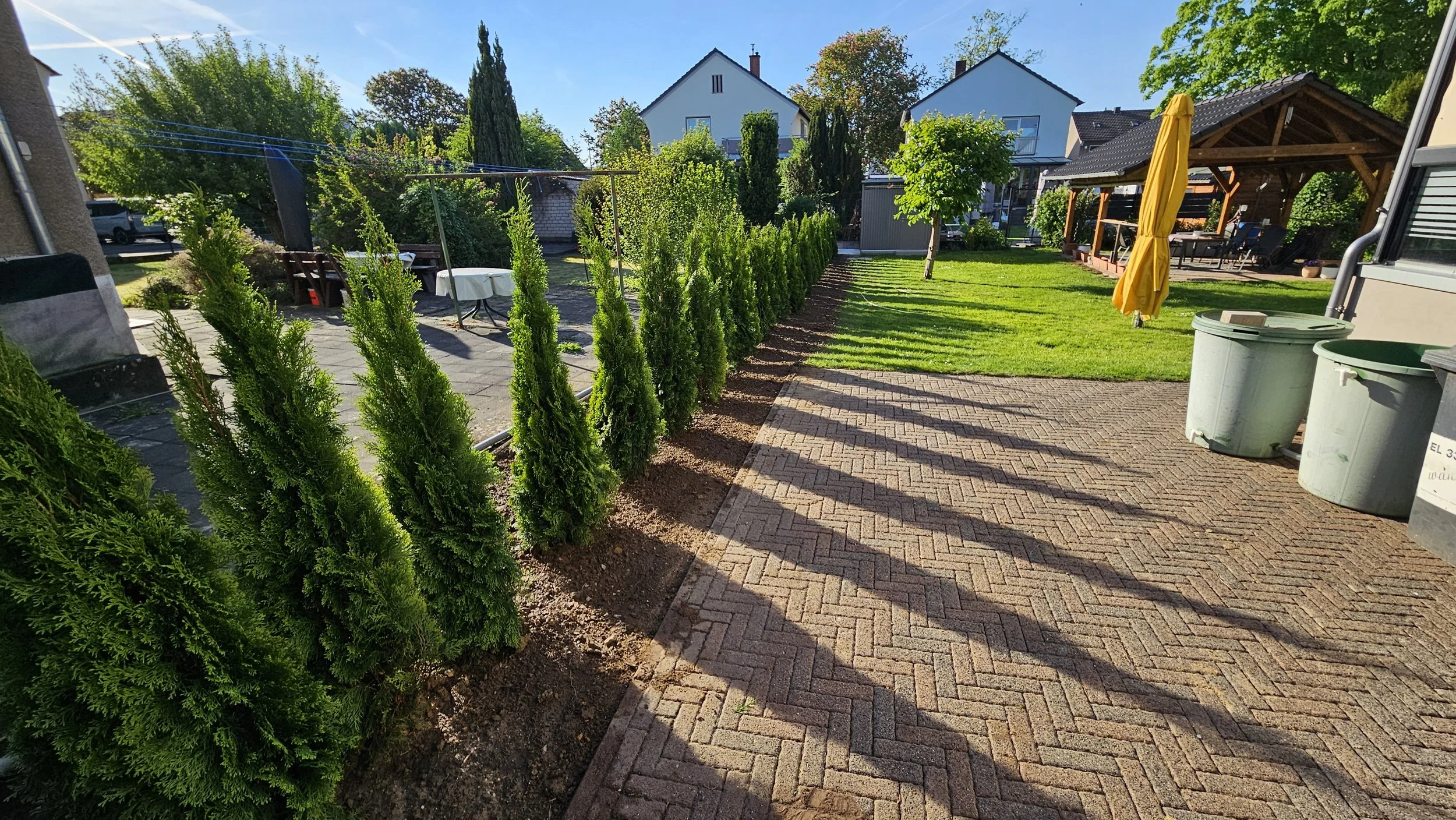 Ein gepflegter Garten mit einer Reihe von hohen, grünen Hecken, einem gepflasterten Weg, einem Rasenbereich, einer Sitzecke mit einem gelben Sonnenschirm, einer Pergola, und mehreren Mülltonnen.