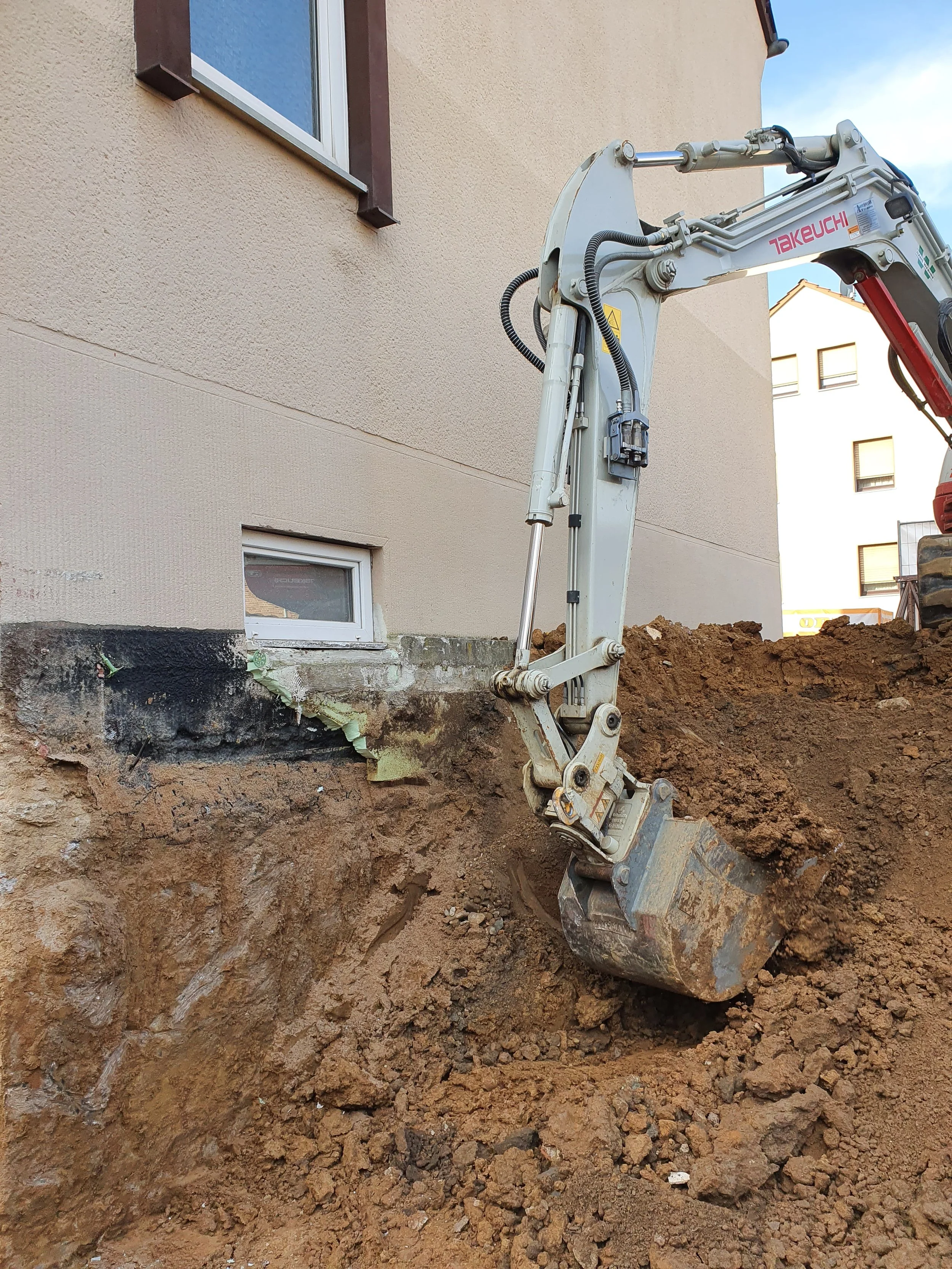 Bauarbeiten an einem Haus, ein Bagger gräbt einen Graben vor der Hauswand.