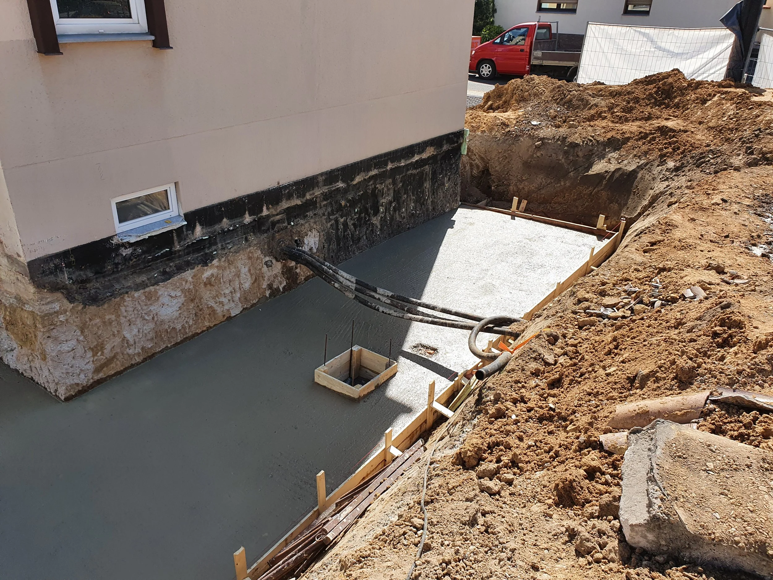 Fundament für eine Terrasse oder einen Balkon wird gegossen, mit Holzrahmen und Eisenstiften, neben einem Haus mit 2 kleinen Fenstern, im Hintergrund Fahrzeuge und Bauzäune.