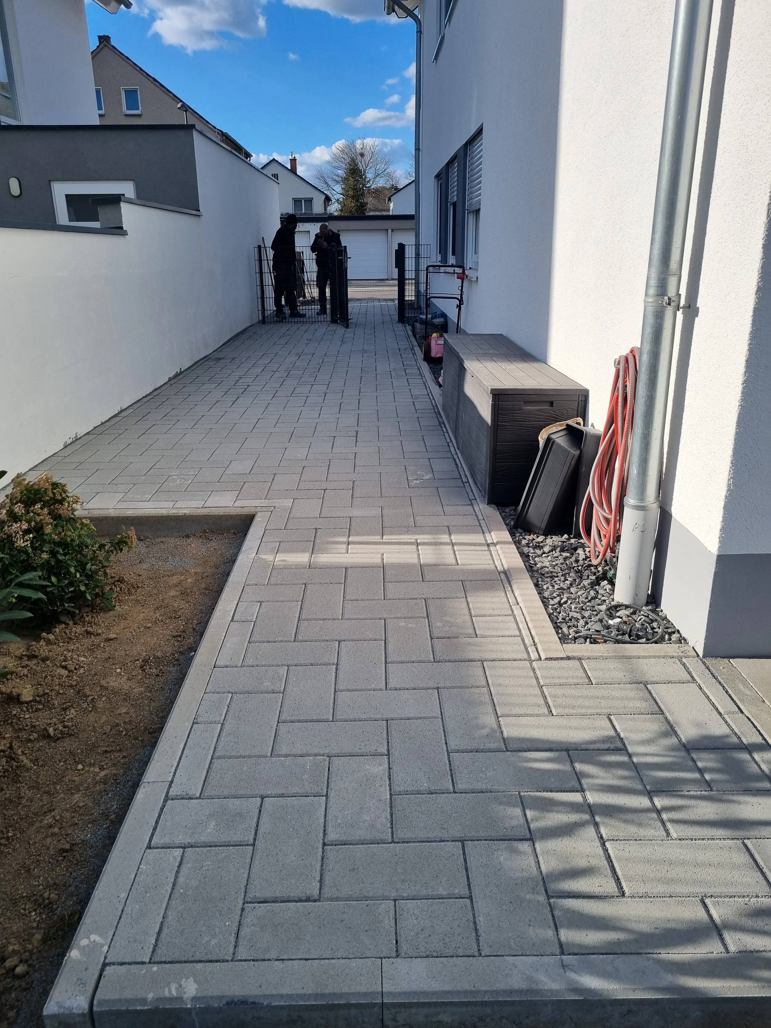 Ein gepflasterter Weg zwischen weißen Hauswänden, mit zwei Männern im Hintergrund, die vor einem Tor stehen.