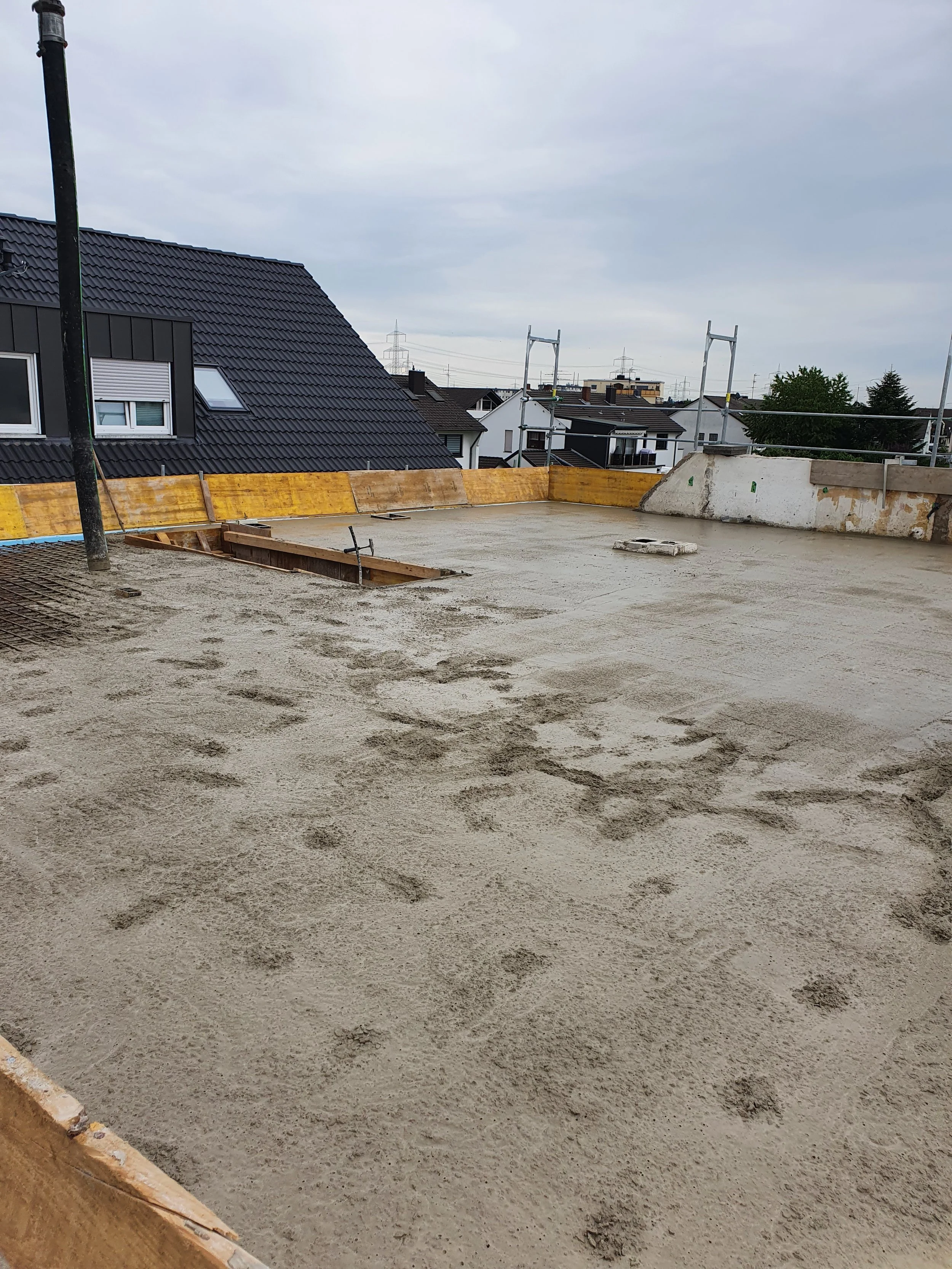 Baugrube auf einer Baustelle mit frisch geglätteter Betonschicht, umgeben von Holzrahmen und in der Nähe eines Hauses mit dunklem Dach.