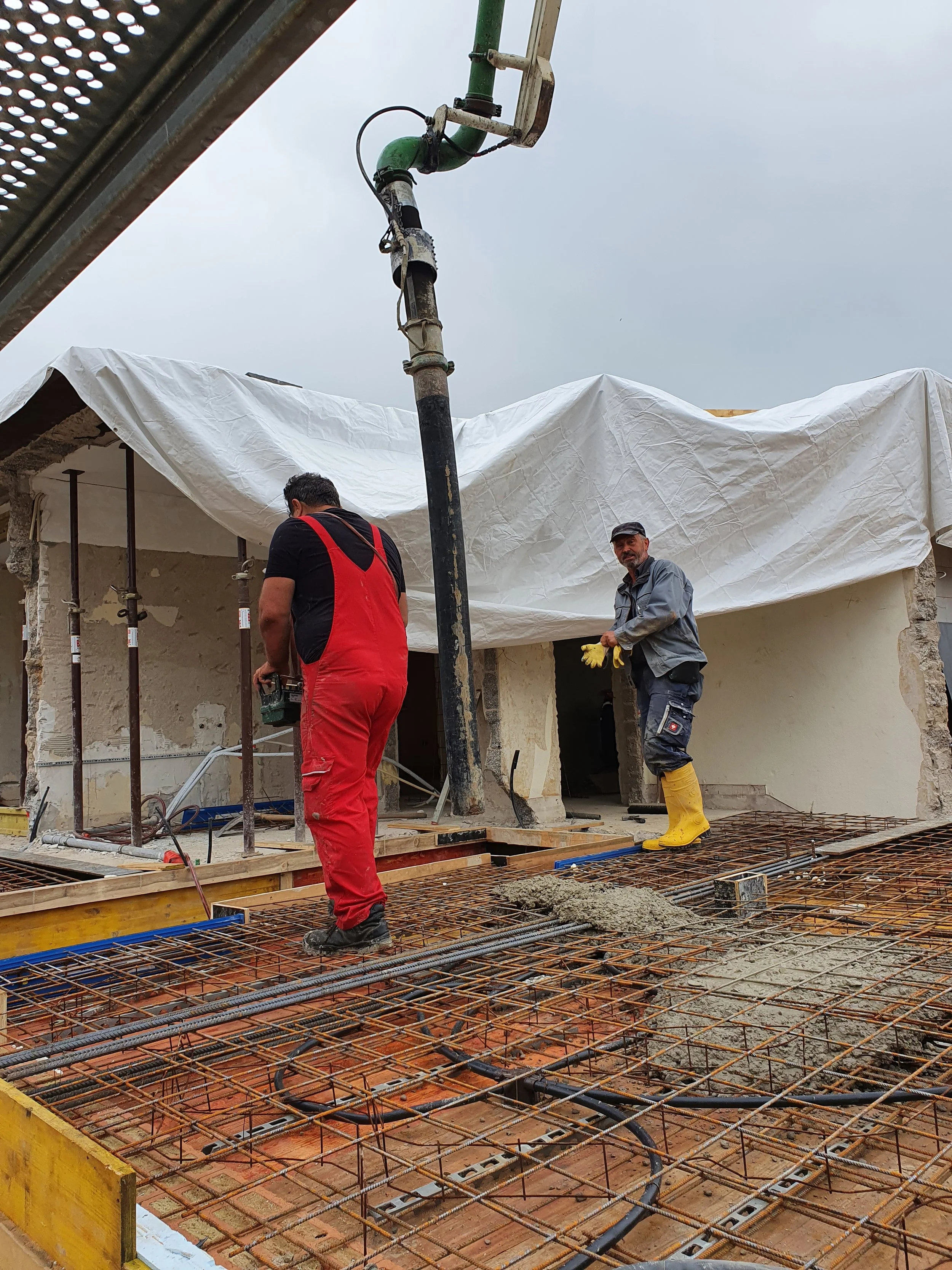 Zwei Bauarbeiter beim Betonieren auf einer Baustelle, mit Stahlbewehrung und Dachkonstruktion im Hintergrund.