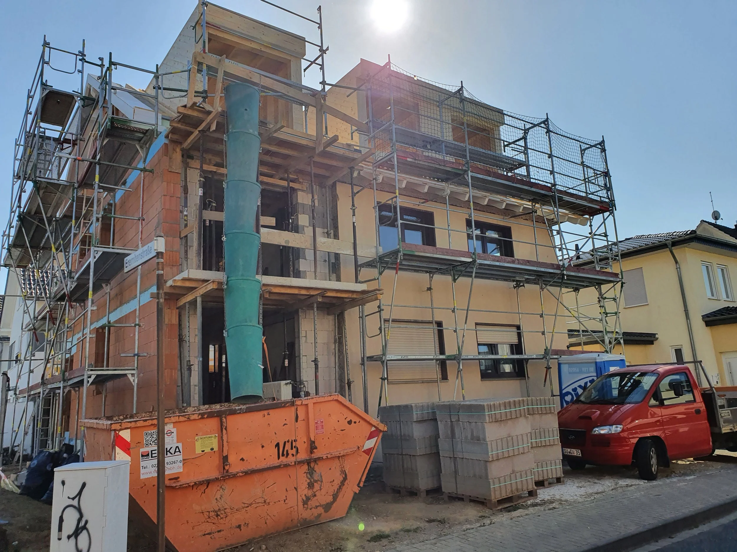 Ein zentrales Mehrfamilienhaus im Bau, eingerüstet mit Baugerüsten, Baucontainer und Baumaterialien vor einem sonnigen Himmel, in einer Wohnsiedlung.