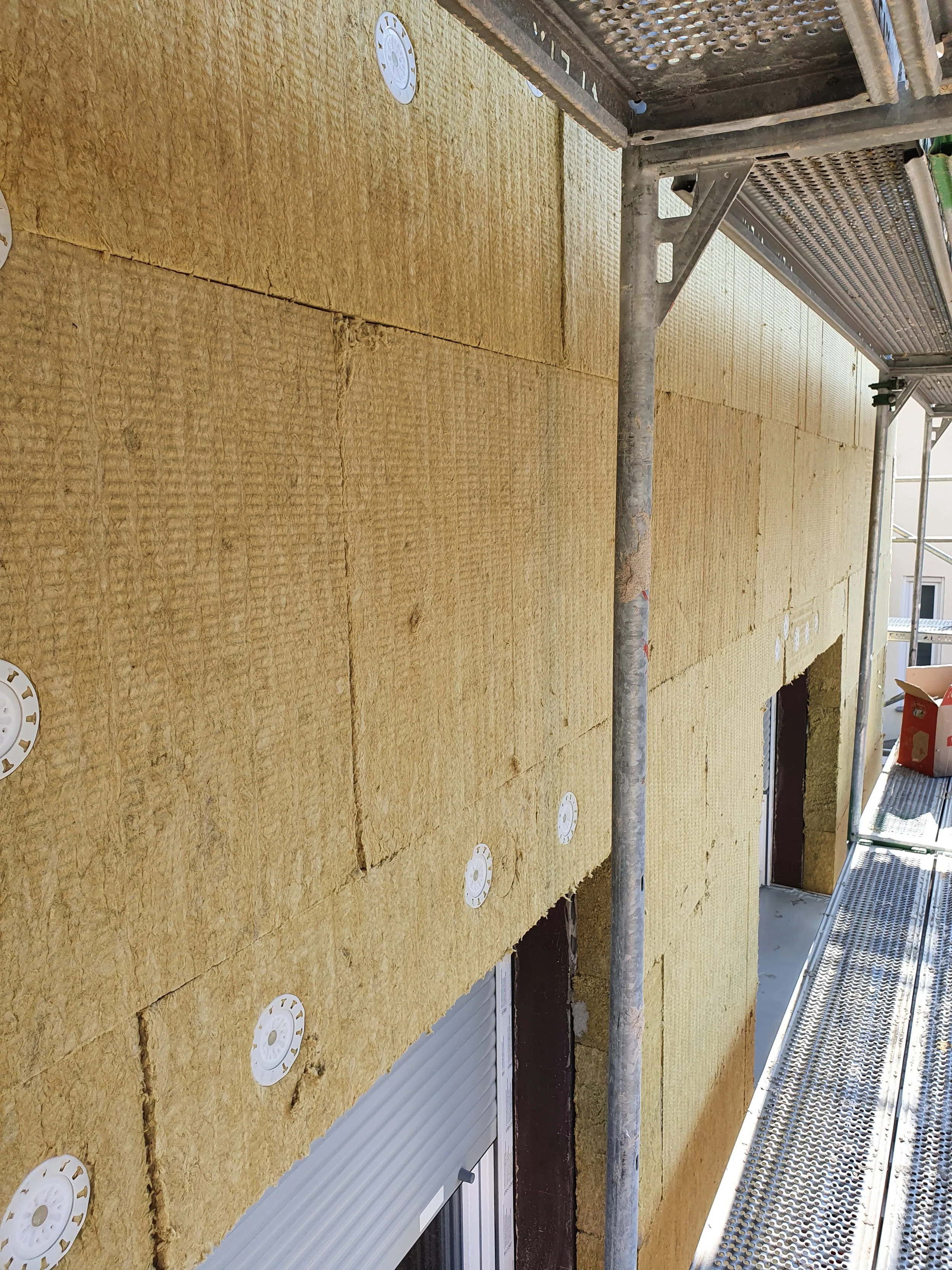 Außenwand eines Gebäudes, das mit Dämmmaterial gedämmt wird, auf einer Baugerüsteile sitzend.