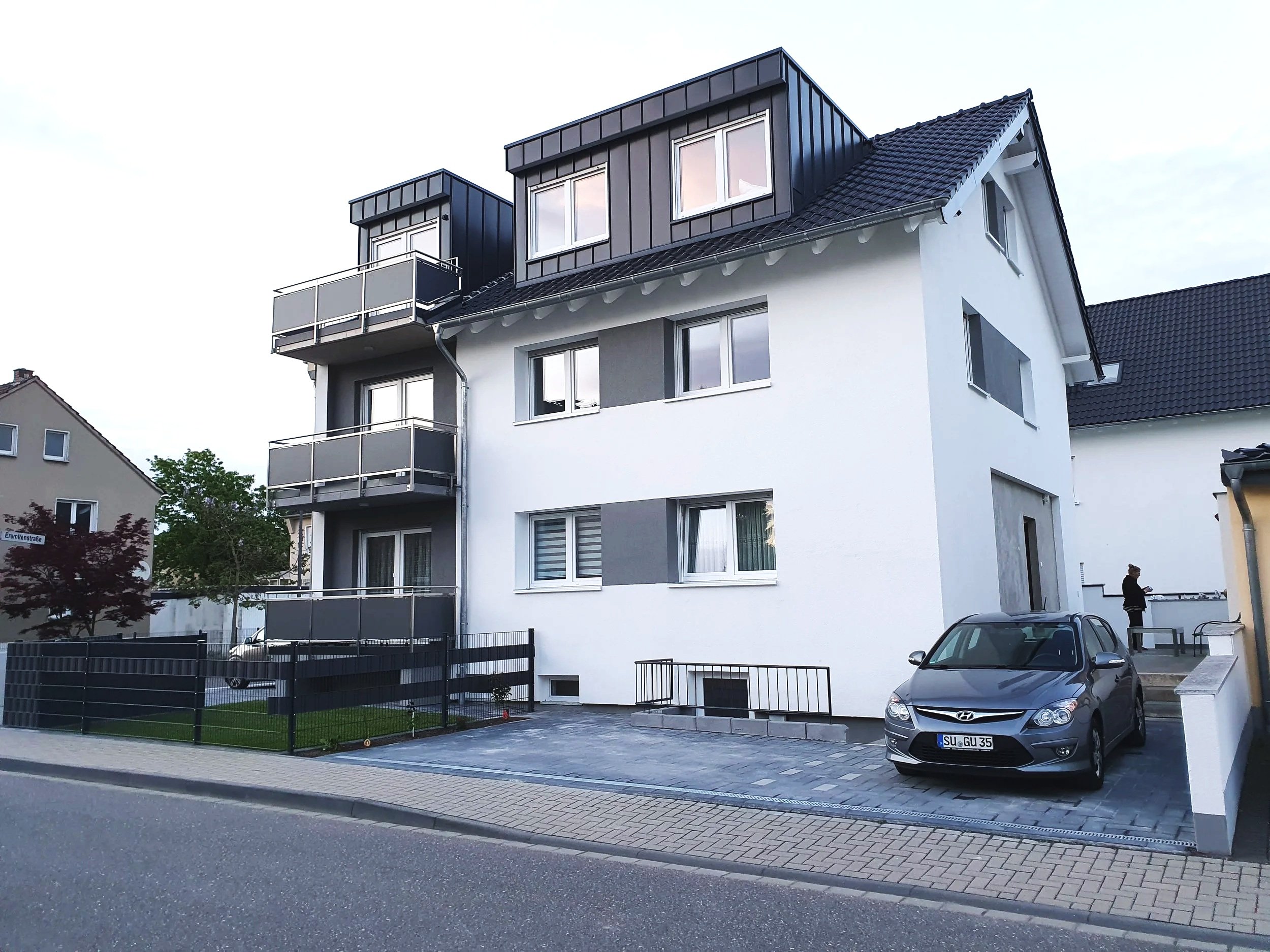 Mehrstöckiges modernes Wohnhaus mit weißen und grauen Wänden, Balkonen und einem Pkw vor dem Gebäude.