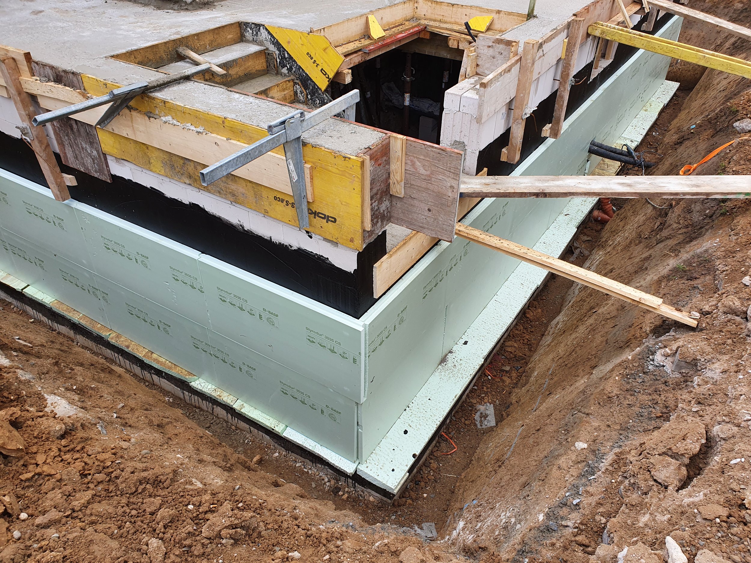 Fundament eines Gebäudes im Bau mit Isolierung, Holzrahmen und Bauwerkzeugen, umgeben von Erdreich.