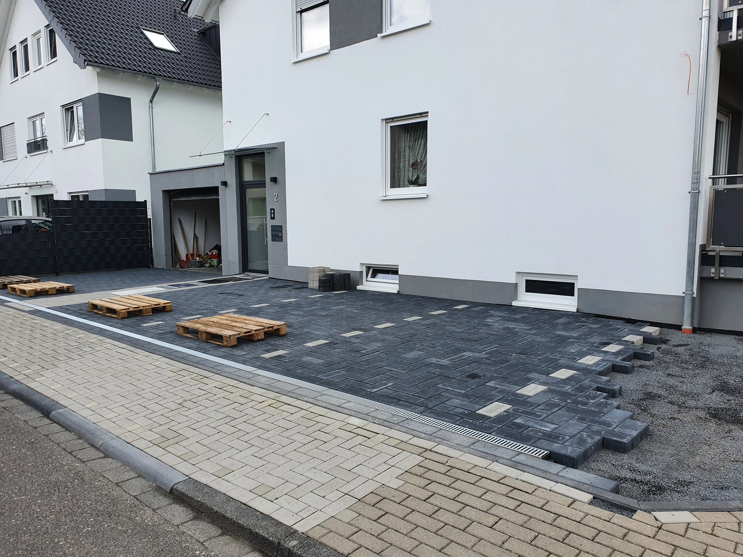 Neuer gepflasterter Parkplatz vor einem weißen Mehrfamilienhaus mit dunklen Pflastersteinen, Paletten mit Pflastersteinen und Gartenwerkzeugen im Kellerbereich.