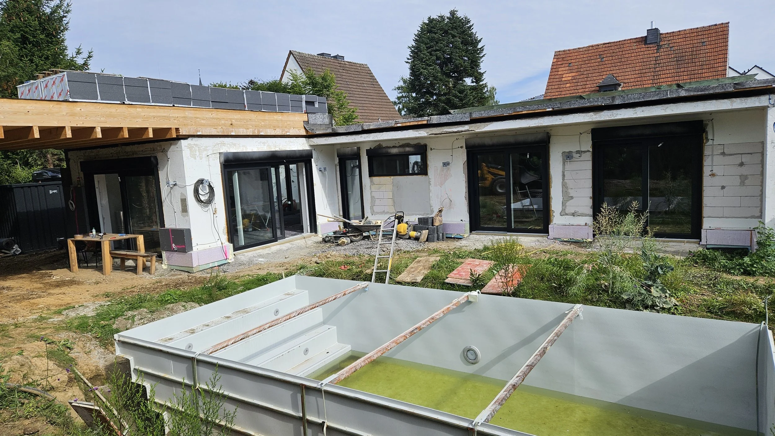 Reges Bauarbeiten an einem modernen Haus mit großen Fenstern, Baugeräten und einem teilweise fertiggestellten Swimmingpool im Vordergrund.