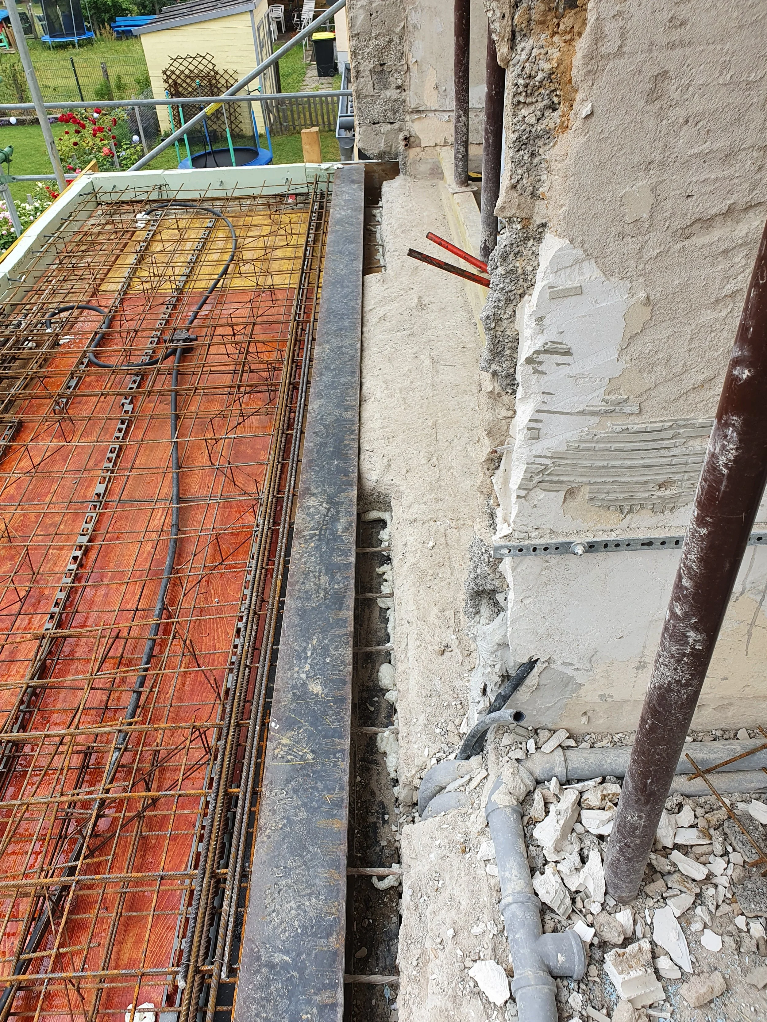 Baustelle mit Stahlarmierungen, Rohren und zerbrochenem Mauerwerk an einer Gebäudefassade, sichtbare Bauarbeiten im Außenbereich.