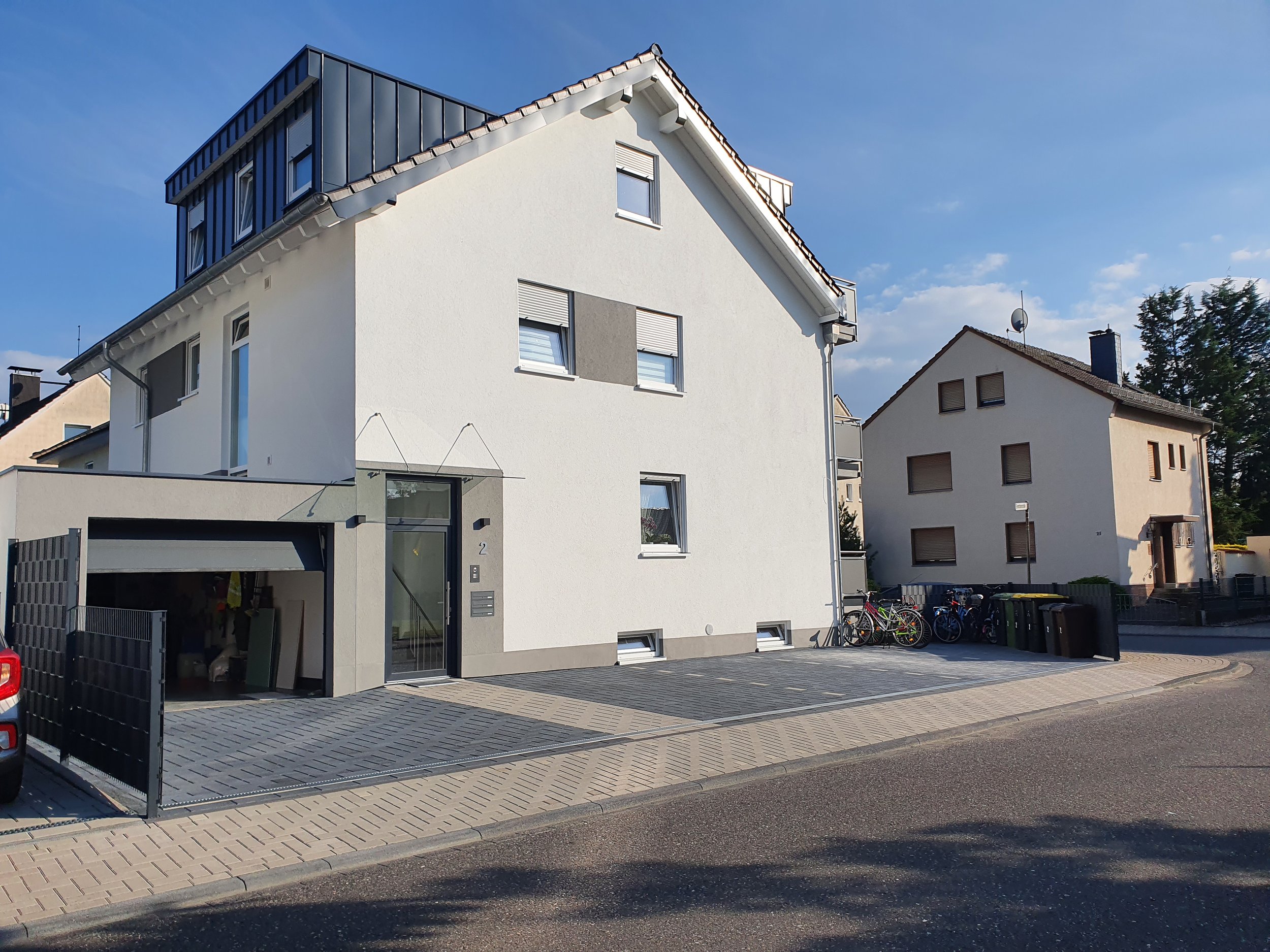 Modernes mehrstöckiges weißes Haus mit Garage, Fahrradständer und Mülltonnen, in einer ruhigen Wohnstraße bei sonnigem Wetter.