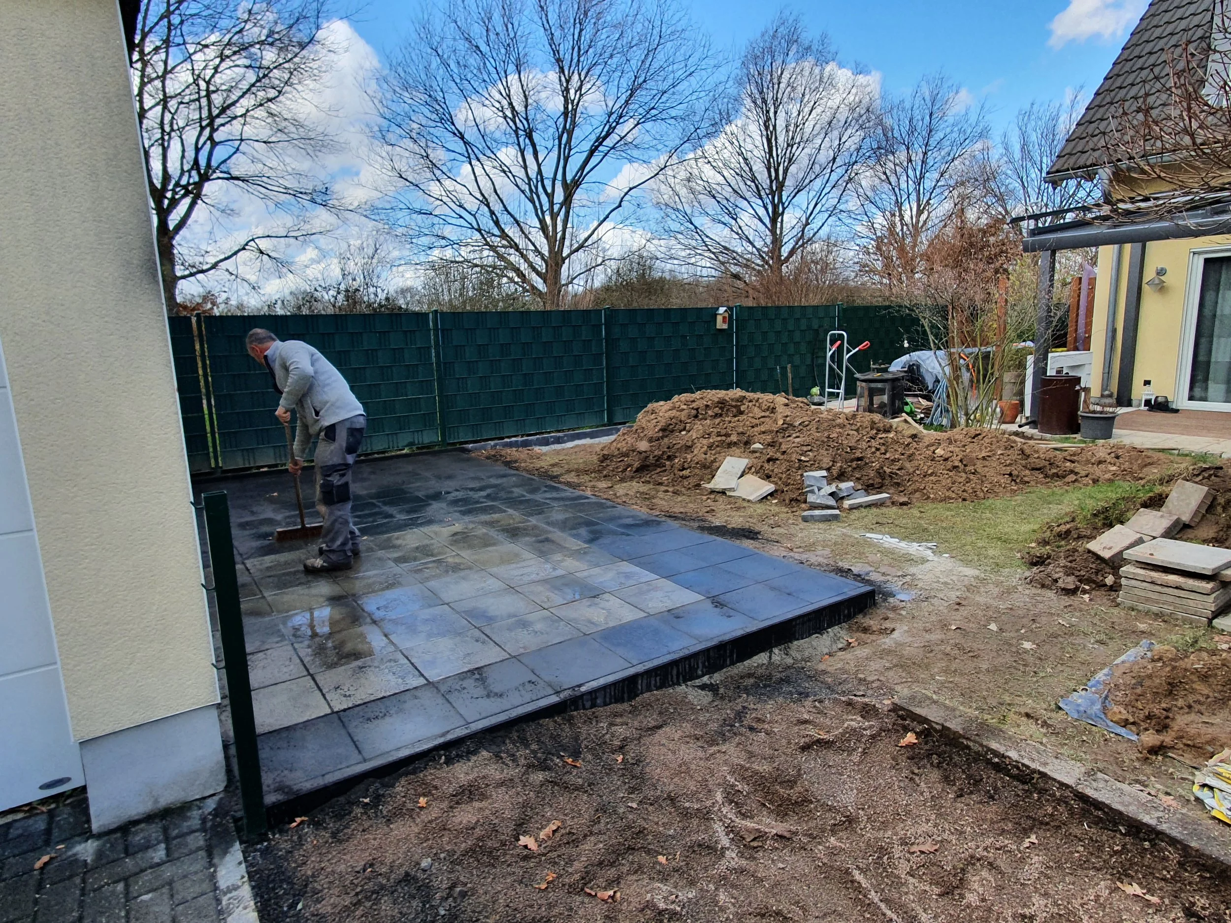 Ein Mann verlegt Pflastersteine auf einer Terrasse vor einem Haus. Im Hintergrund sind Bauarbeiten und Erde zu sehen, sowie ein grüner Zaun und Bäume ohne Blätter.