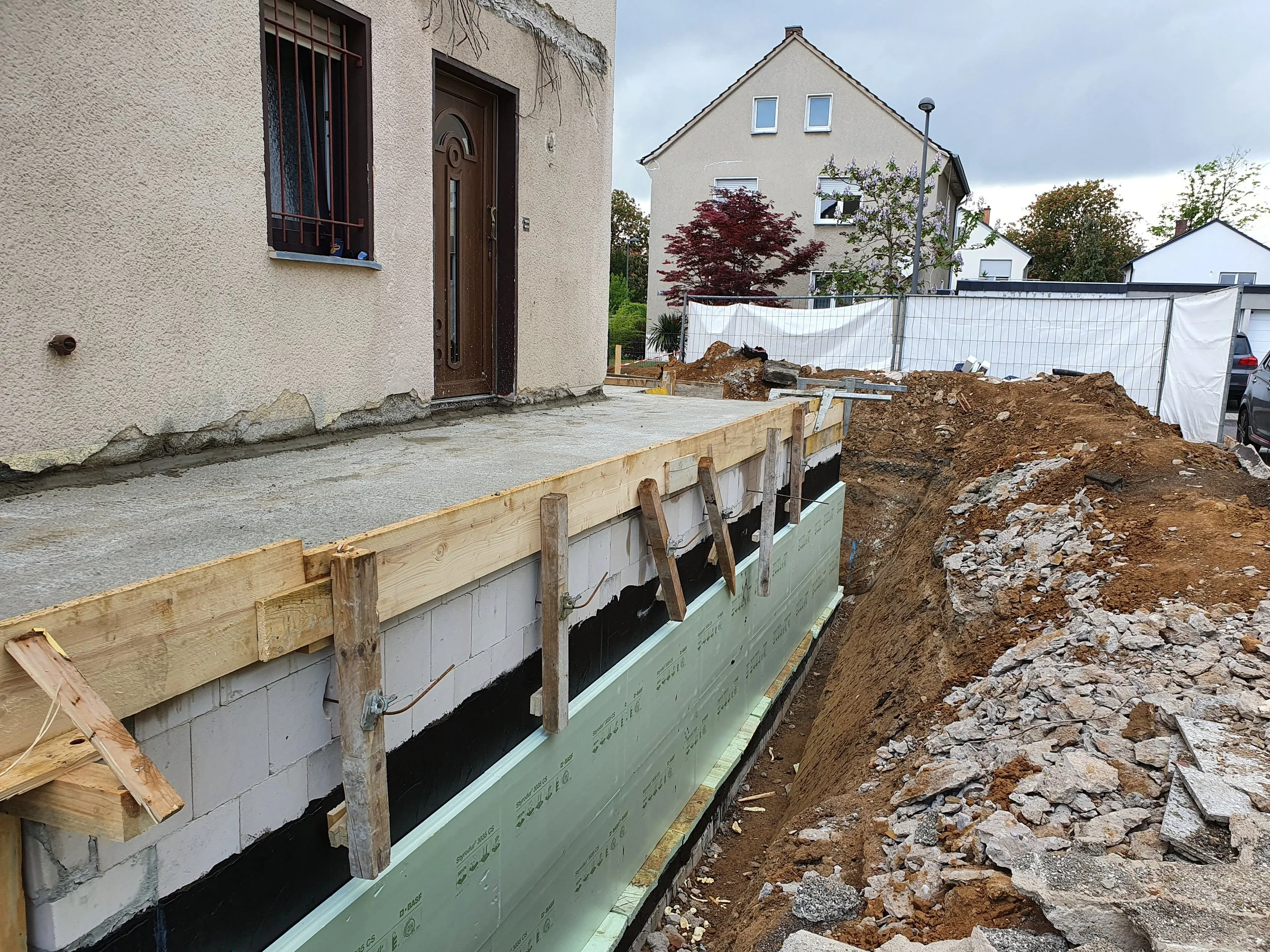 Bauarbeiten an Hausfundamenten mit Bauholz, Erde und Steinen gespeichert. Das Haus hat eine beige Wand und eine Tür, im Hintergrund sind andere Häuser und Bäume sichtbar.
