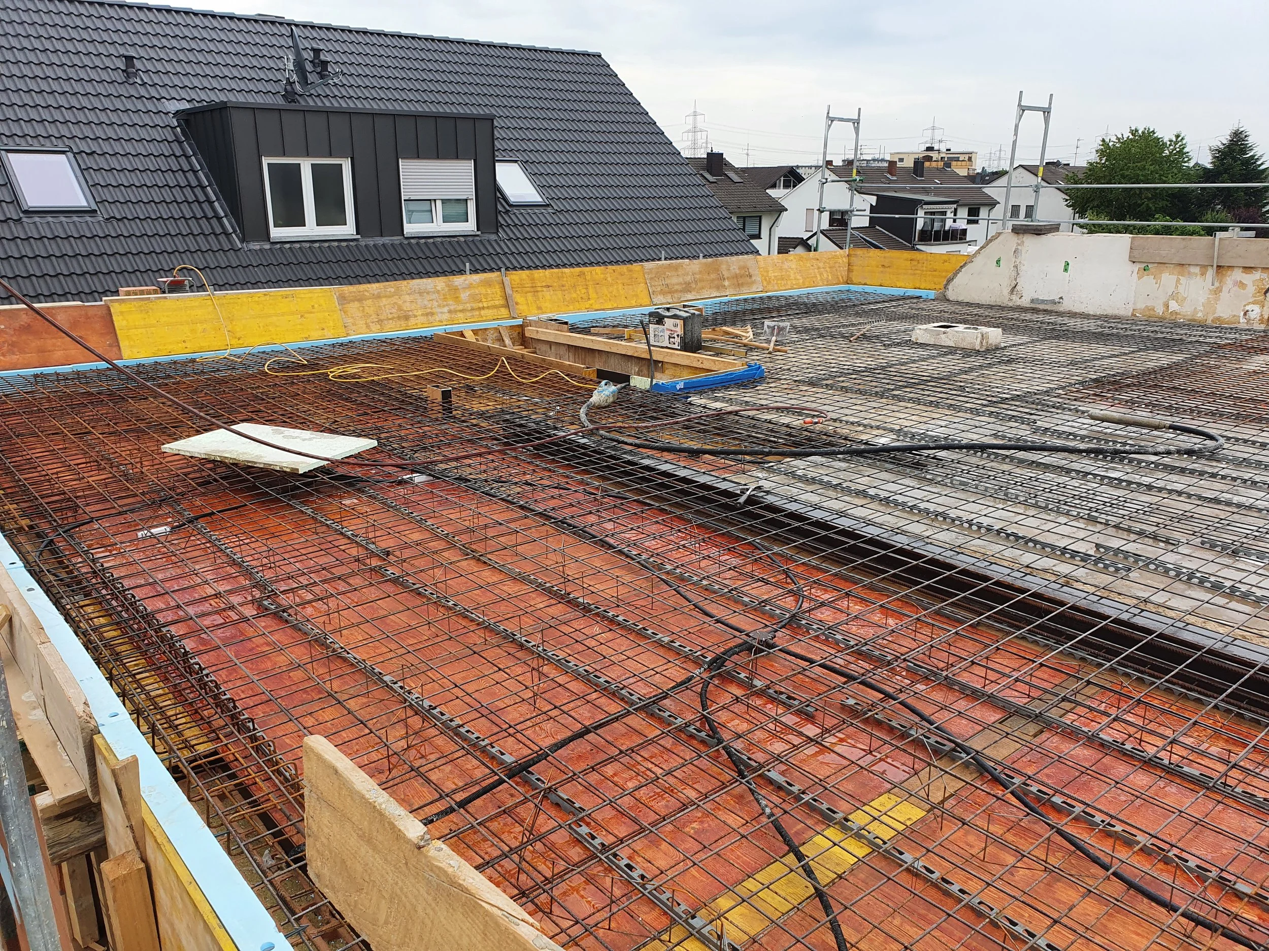 Baustelle mit Eisenbewehrung für den Deckenbau auf einem Dach, im Hintergrund Häuser und ein bewölkter Himmel.