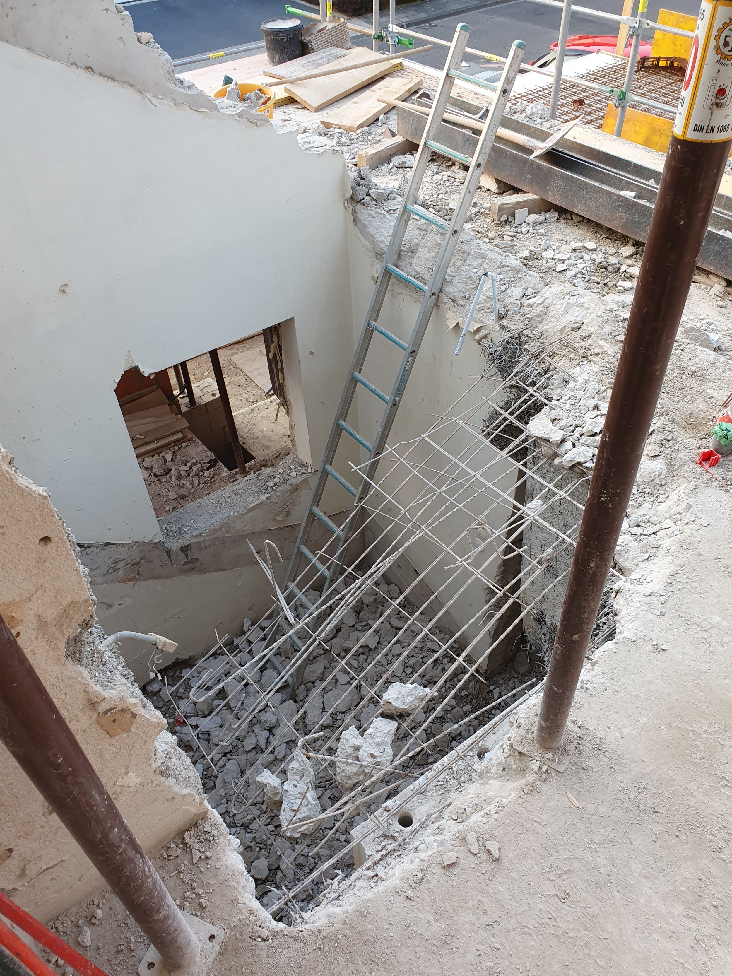 Baustelle mit ausgeschnittenem Boden, eine Leiter, Stahlbewehrung und teilweise abgerissene Wand.