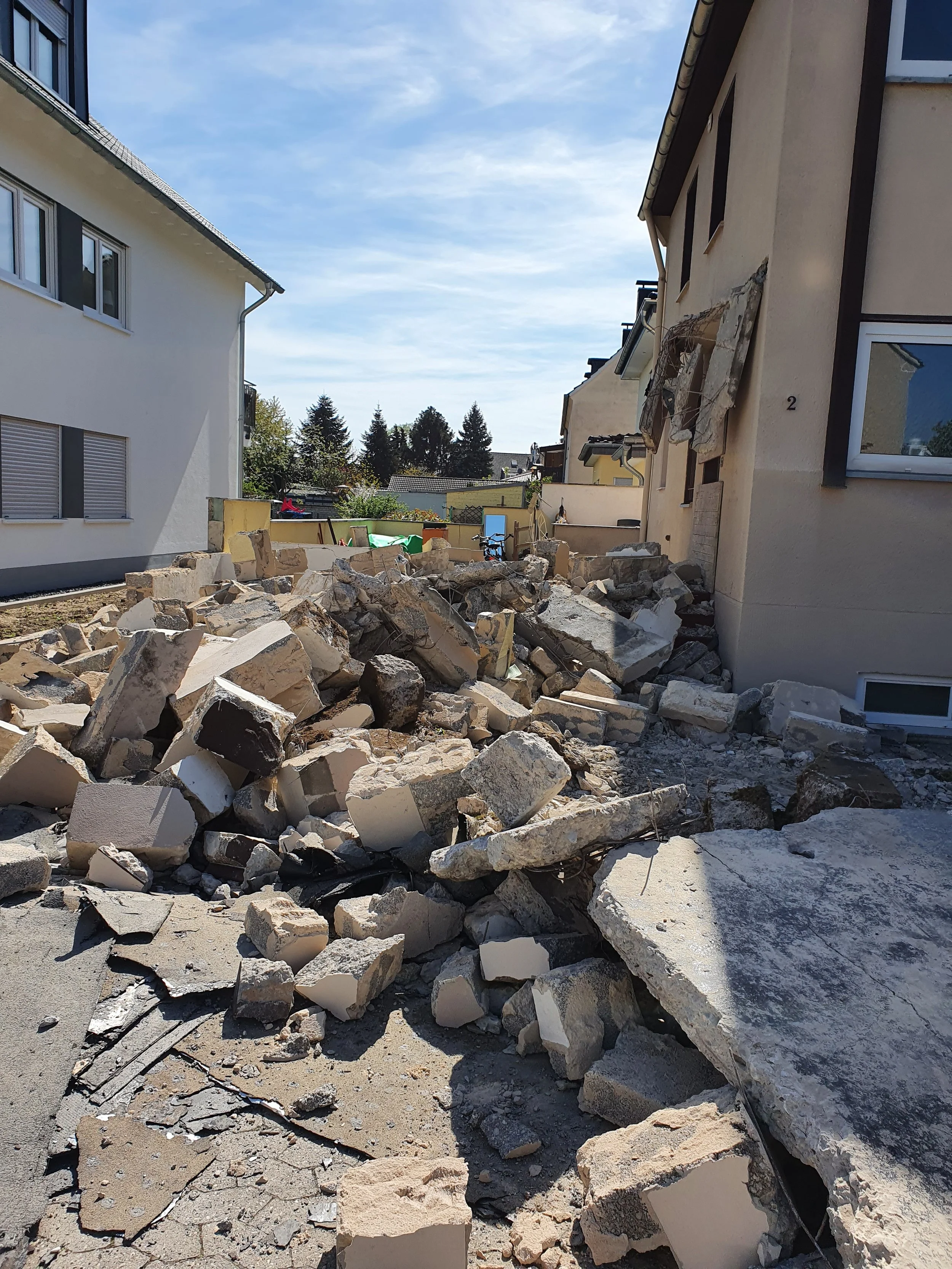Zerstörte Wand und Trümmer auf einem Gehweg vor einem Haus, verursacht vermutlich durch einen Schaden oder Abriss.