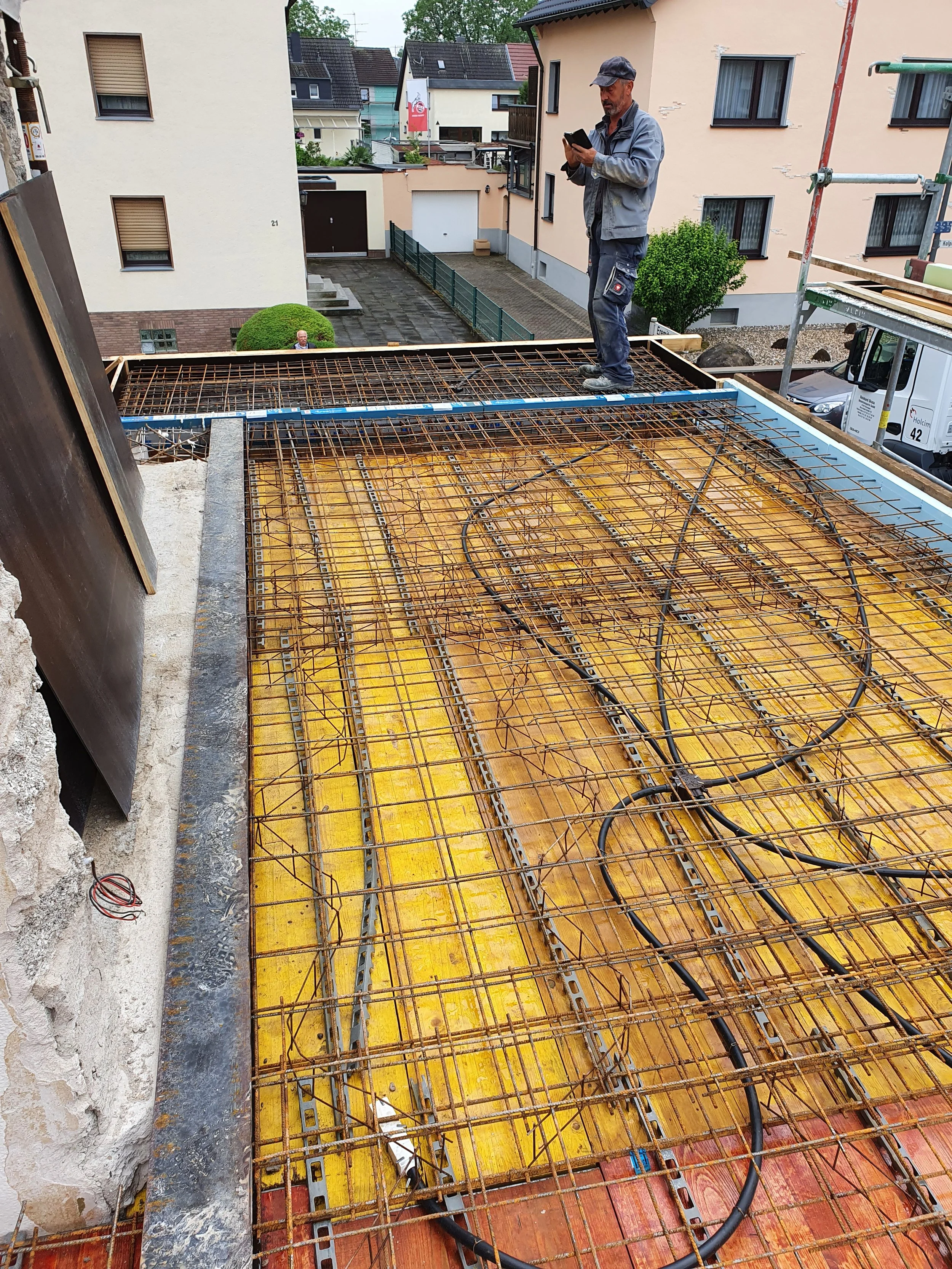 Bauarbeiter steht auf einem Baugerüst und sieht auf sein Mobiltelefon. Im Vordergrund sind Stahlverstärkungen und Kabel auf einer Baustelle sichtbar.