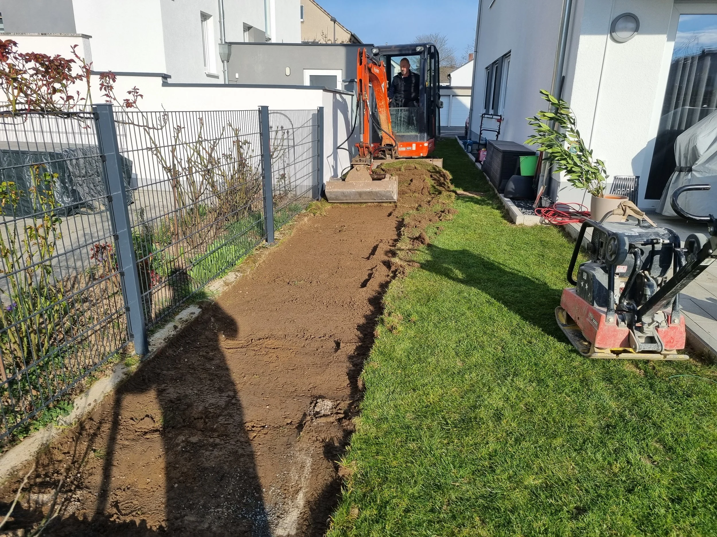 Gartenarbeit beim Terrain ausheben mit einem Minibagger neben einer weißen Hauswand, auf der Gartengeräte und Pflanzen stehen, während im Hintergrund ein Gebüsch und andere Häuser sichtbar sind.