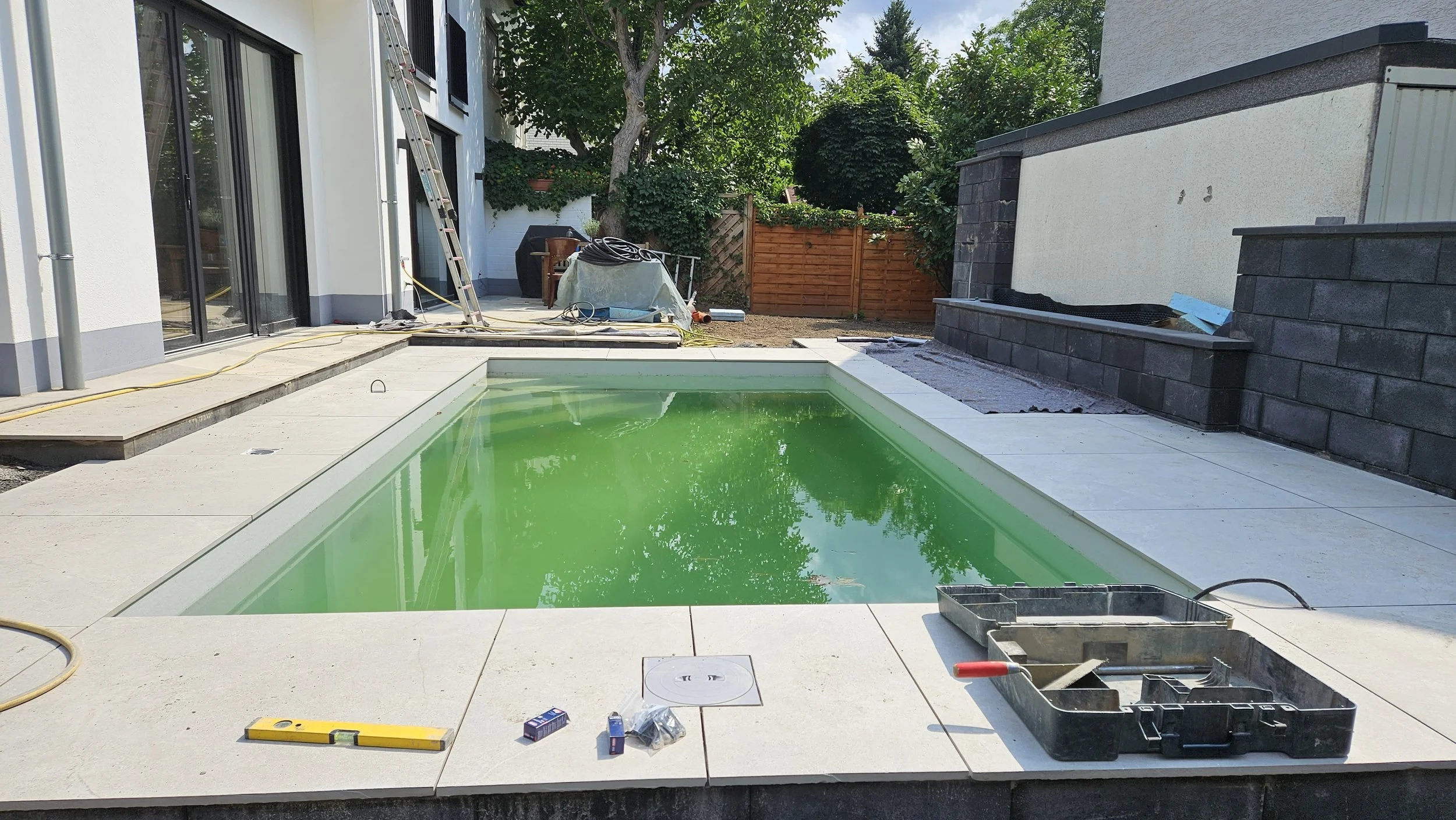 Im Garten eines Hauses wird ein kleiner Swimmingpool mit grünem Wasser gebaut oder renoviert. Im Hintergrund sind Bäume, eine Socke, Werkzeuge und Baustoffe sichtbar. Der Bereich ist noch im Bau, es gibt Arbeitstools und Baustoffreste.