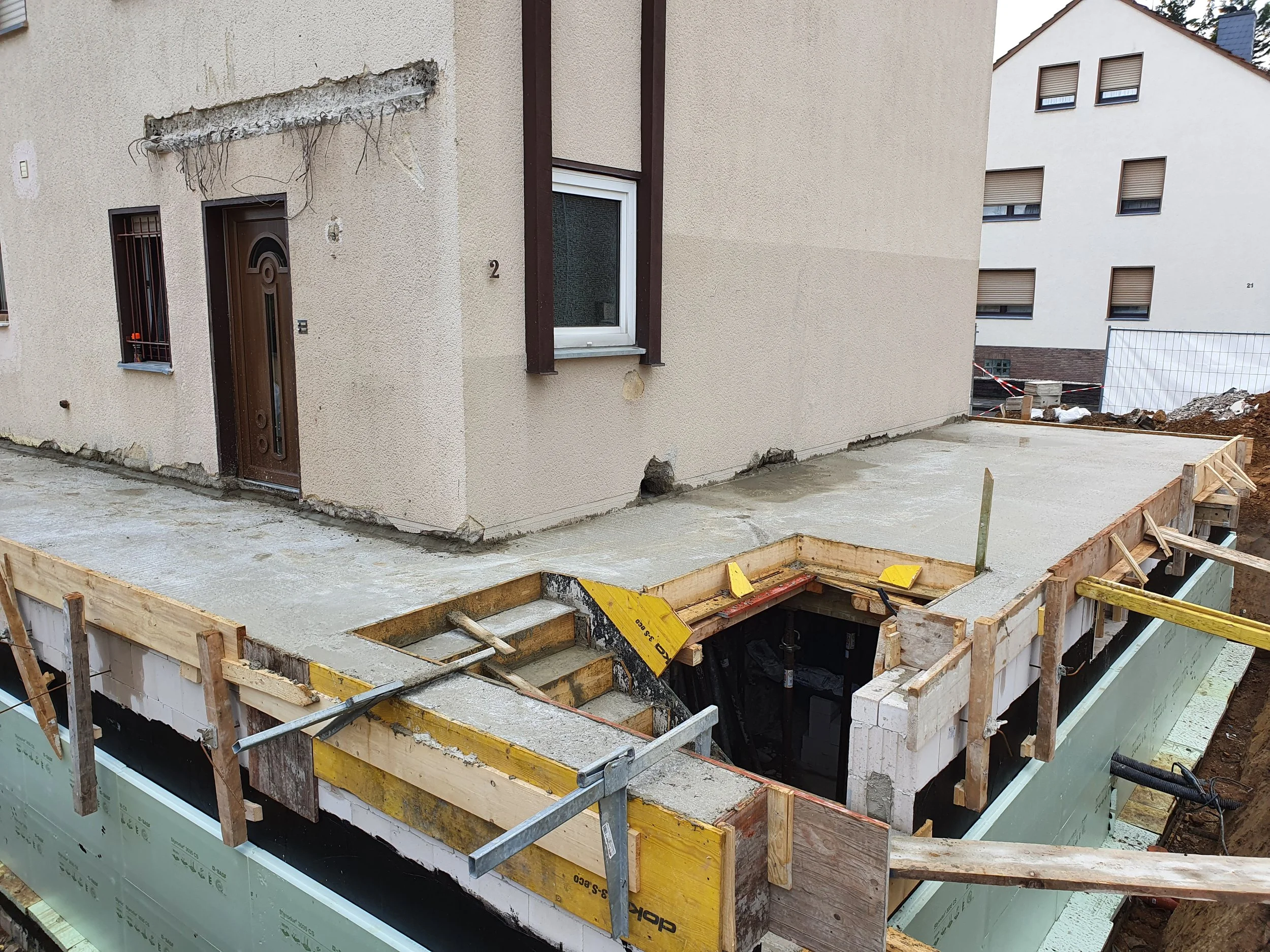 Baustelle vor einem Mehrfamilienhaus mit frisch gegossenem Betonboden, im Vordergrund eine offene Grube mit Holzrahmen für eine Kellertreppe, im Hintergrund einige weitere Mehrfamilienhäuser.