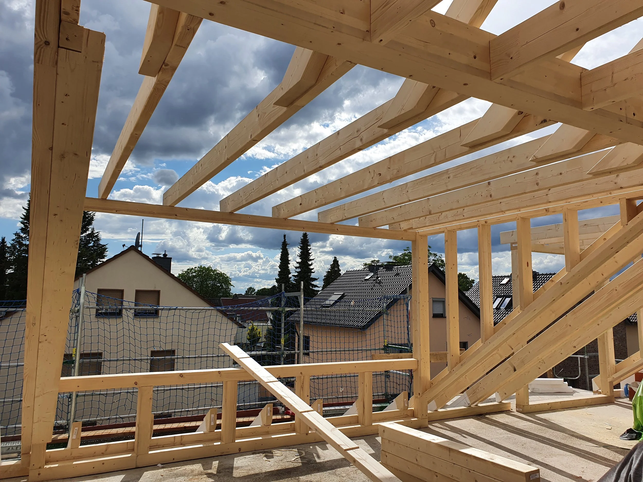 Holzrahmenbau eines Gebäudes unter Bau, mit Dachkonstruktion, umgeben von Nachbarhäusern und bewölktem Himmel.