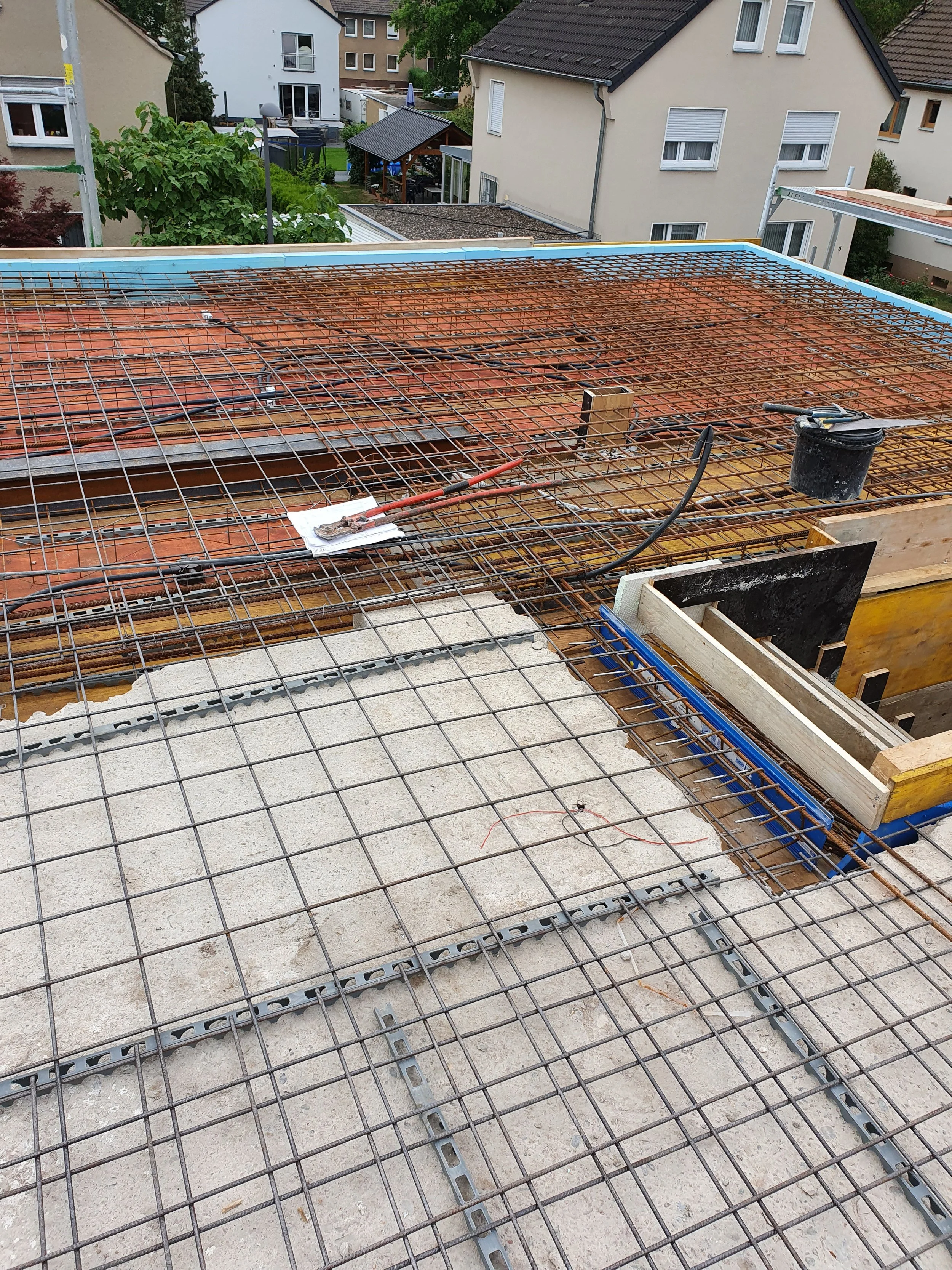 Bauarbeiten auf einem Dach mit Stahlarmierungsnetzen und Bauteilen, im Hintergrund sind Wohnhäuser und Bäume sichtbar.
