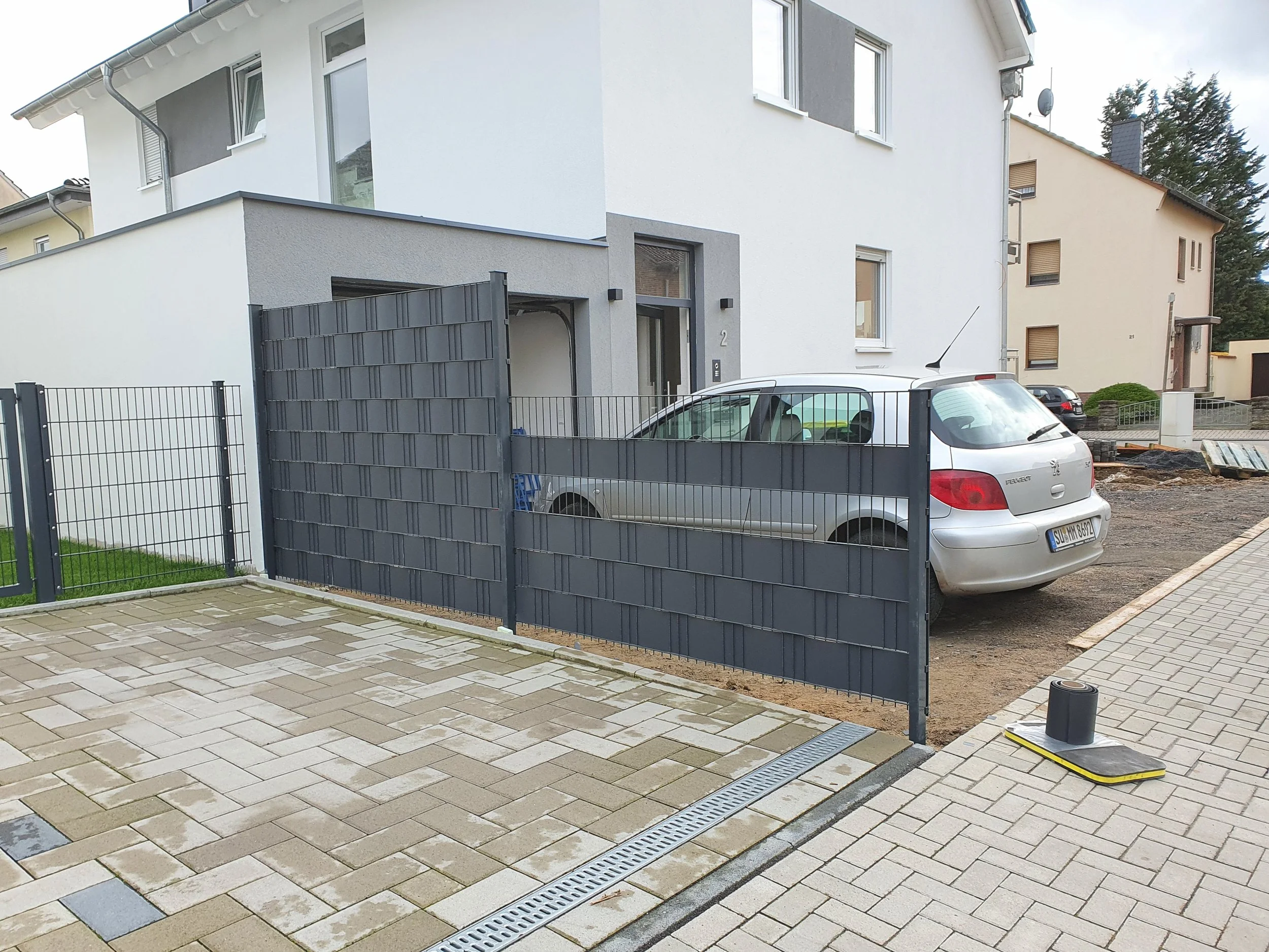 Ein Wohnhaus mit einem geöffneten schwarzen Metallzauntor, darin geparkt ein silberner Peugeot 307. Der Bereich vor dem Haus ist teilweise gepflastert, und rechts liegt ein Pkw-Deckel. Im Hintergrund sieht man weitere Häuser und Bäume.