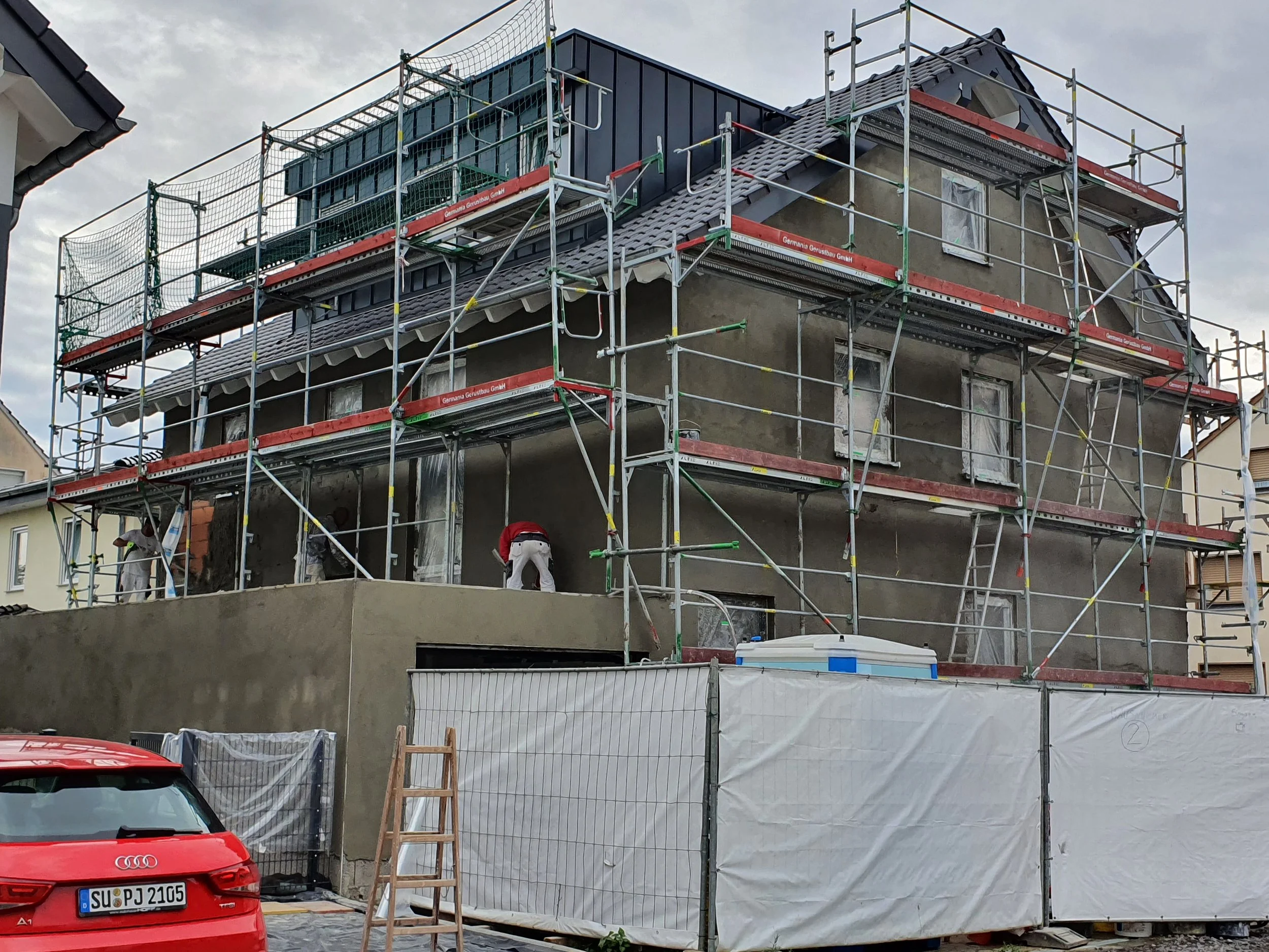 Ein Haus wird renoviert, umgeben von Gerüsten. Es sind Baumaterialien und Arbeiter auf der Baustelle sichtbar.