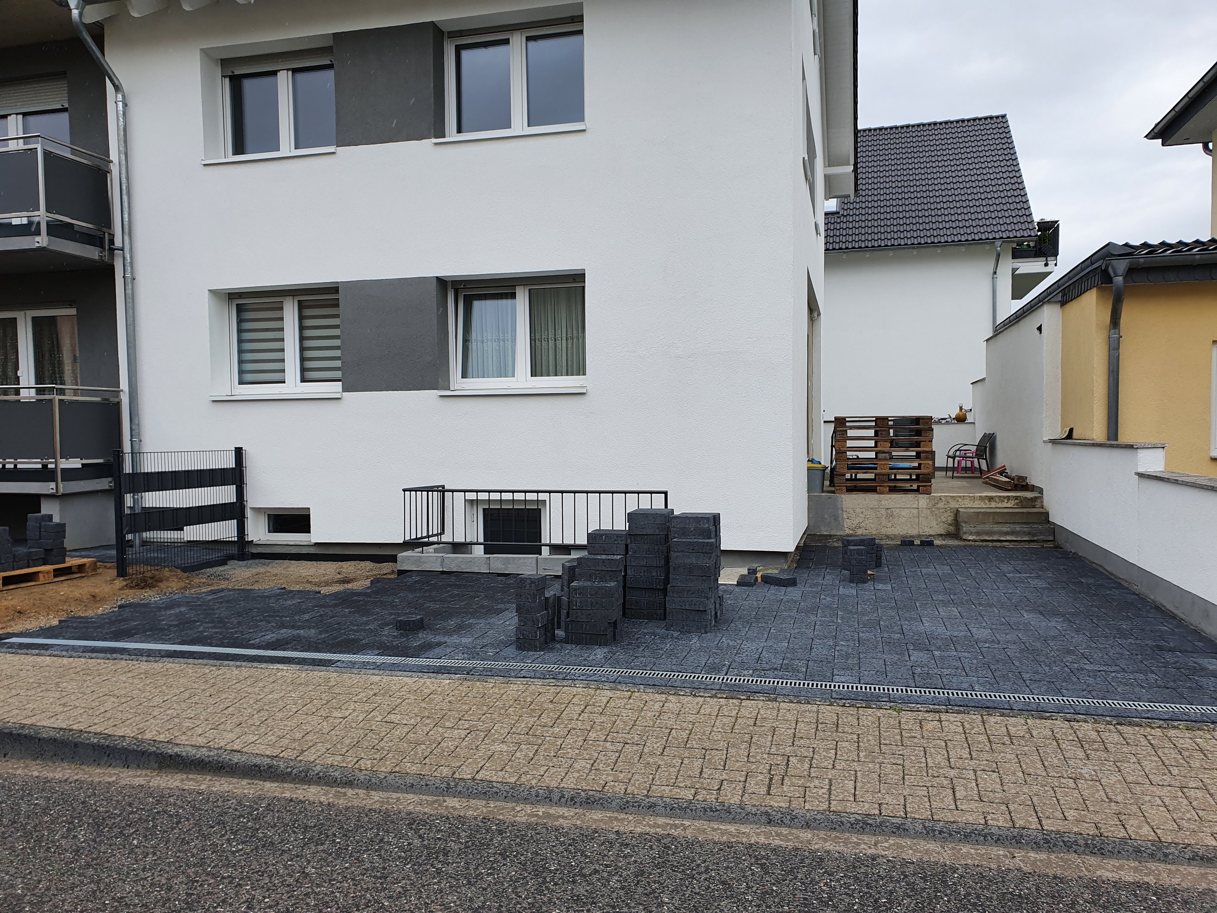 Ein Haus mit einer neuen gepflasterten Terrasse und Baustoffen, Holzplatten, Steine. Das Haus hat weiße und graue Fassaden, mehrere Fenster, eine Treppe und befindet sich in einer Wohngegend mit anderen Häusern.
