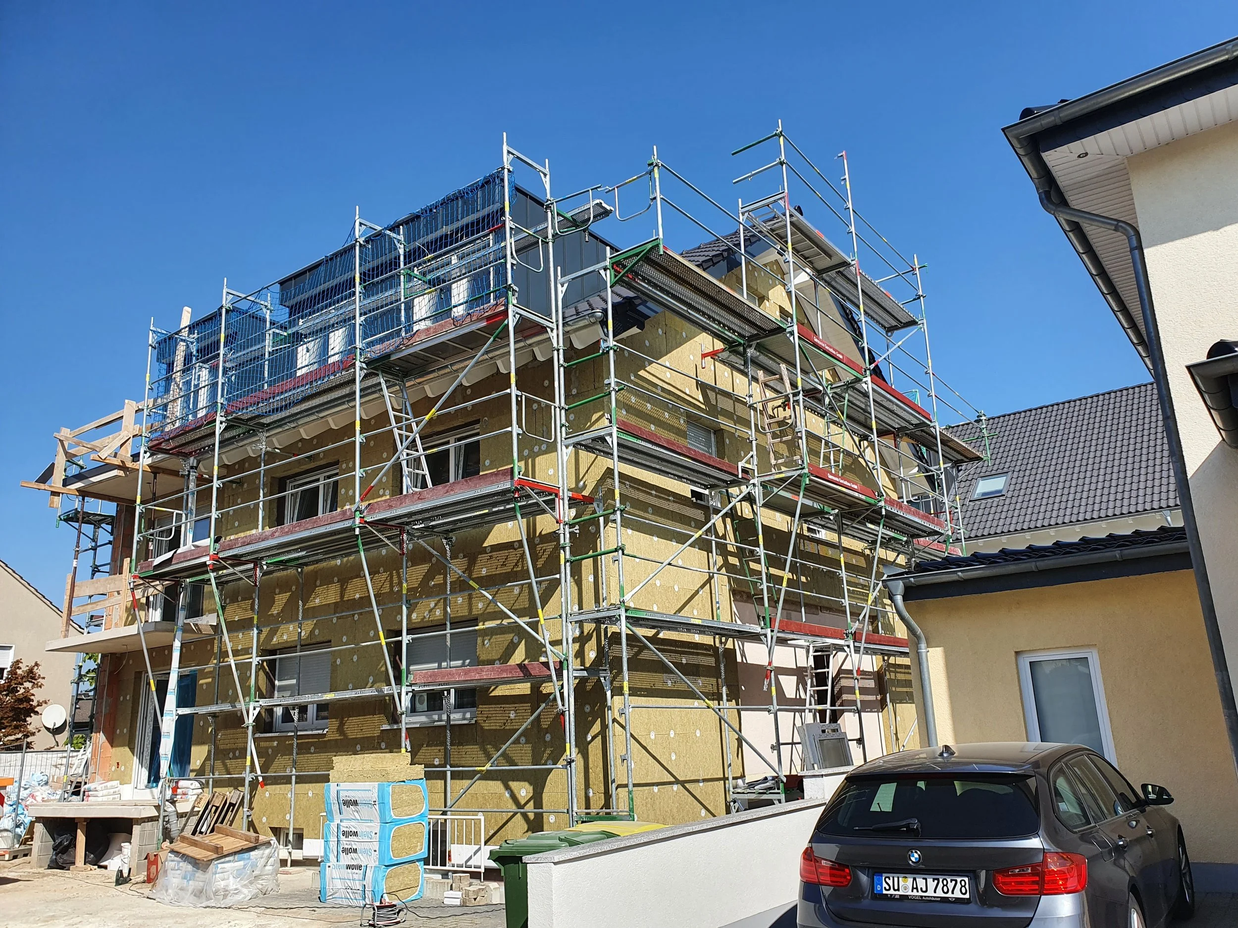 Mehrstöckiges Baugerüst um einen mehrstöckigen Neubau unter Sanierung, Bauarbeitsmaterialien im Vordergrund, ein schwarzer BMW, blauer Himmel.