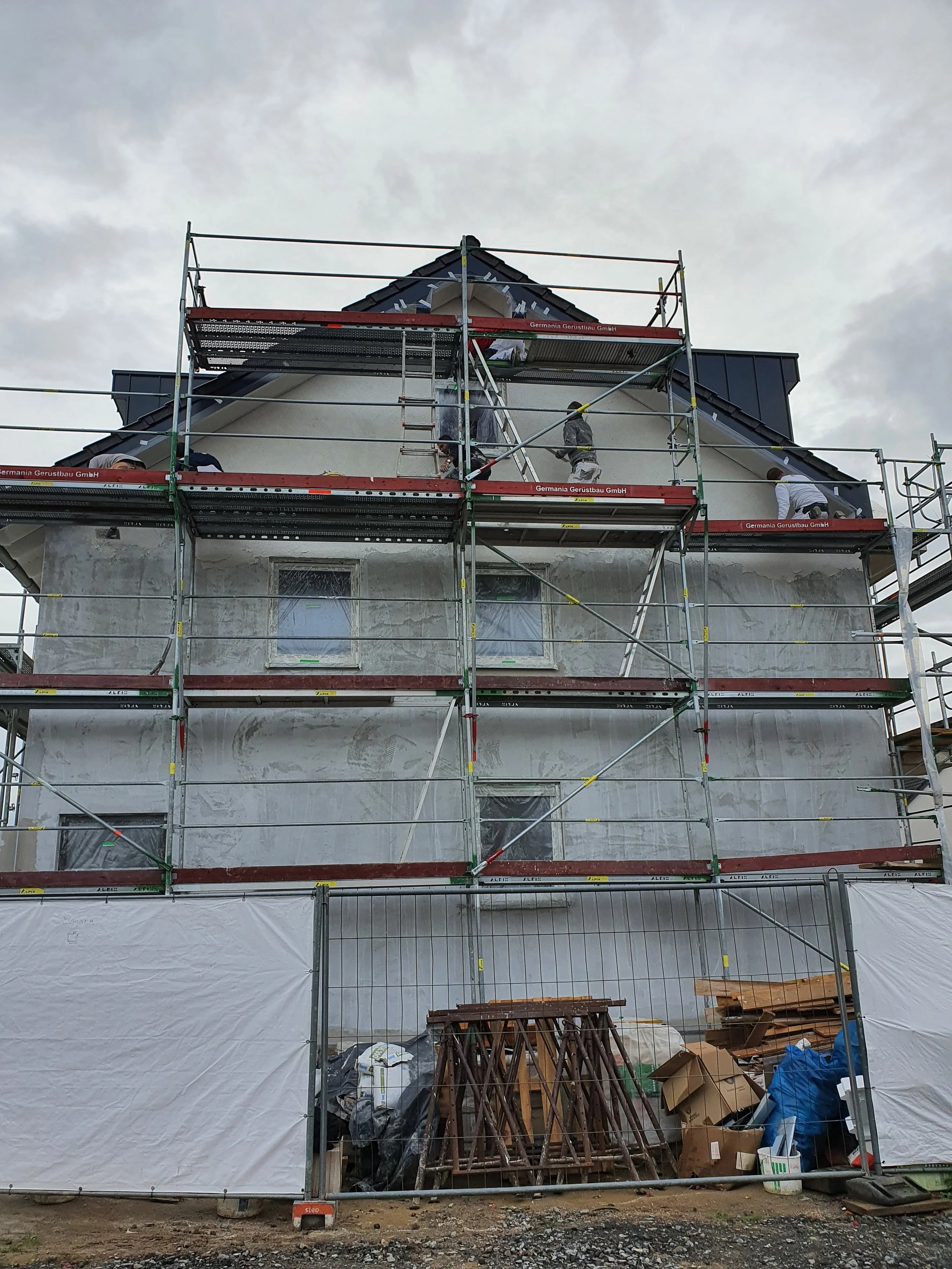 Ein Haus, das gerade renoviert wird, mit Gerüst umgeben, Arbeiter auf dem Dach arbeiten, Wolkenhimmel im Hintergrund.