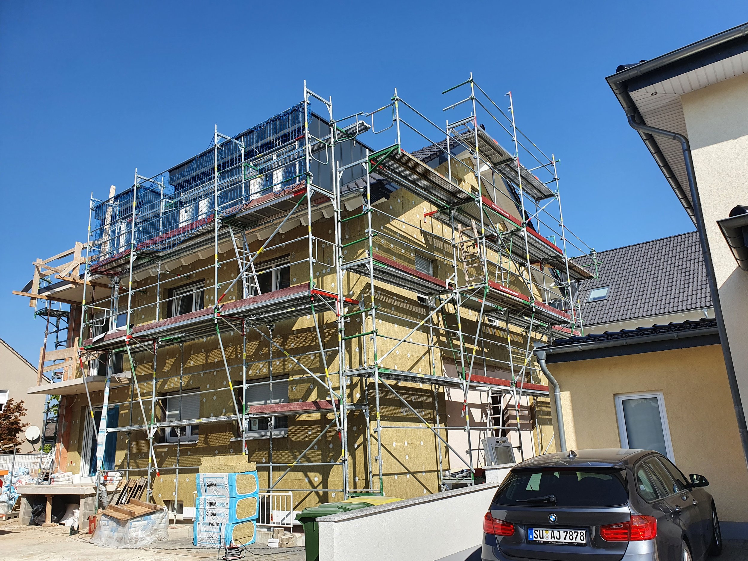 Mehrfamilienhaus unter Sanierung mit Gerüsten, Bauarbeiten an der Fassade, vor einem blauen Himmel, daneben ein schwarzer BMW