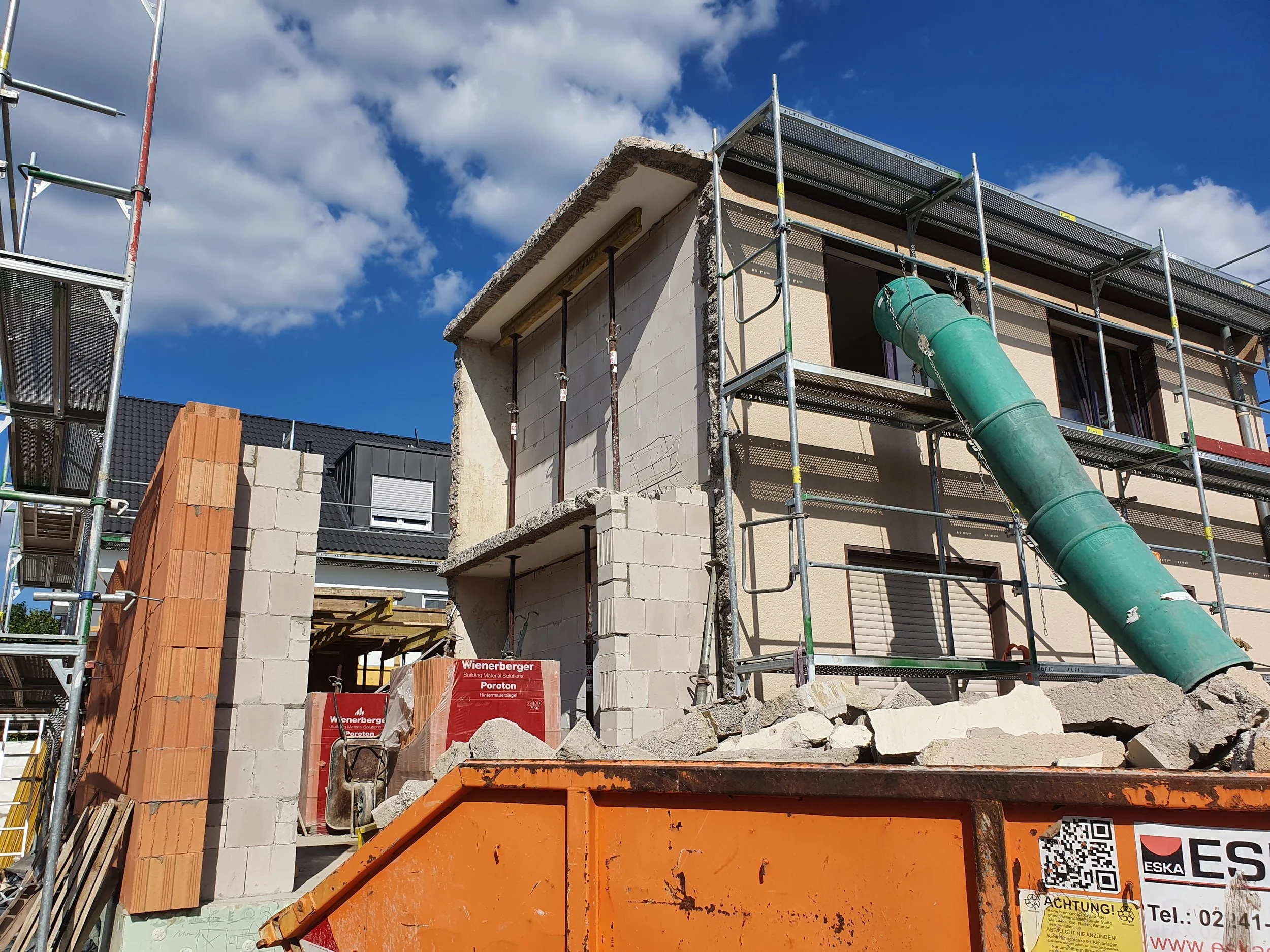 Bauarbeiten an einem Mehrfamilienhaus mit Baugerüst, grünem Rutschtunnel und Baumaterialien im Vordergrund bei schönem Himmel.