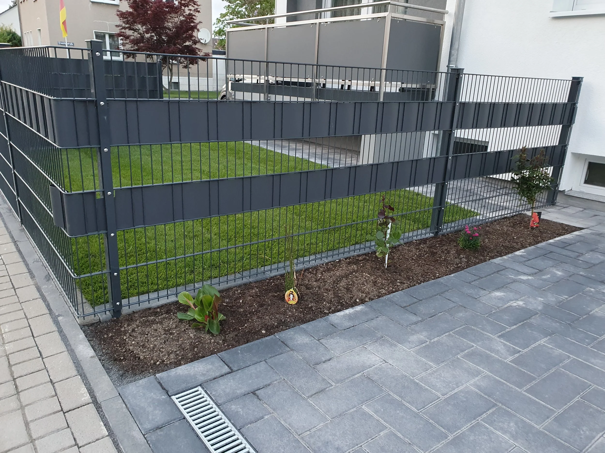 Eingezäunte kleine Gartenfläche mit jungen Pflanzen und Blumen, umgeben von Gehweg und Plattenboden in einem Wohngebiet.
