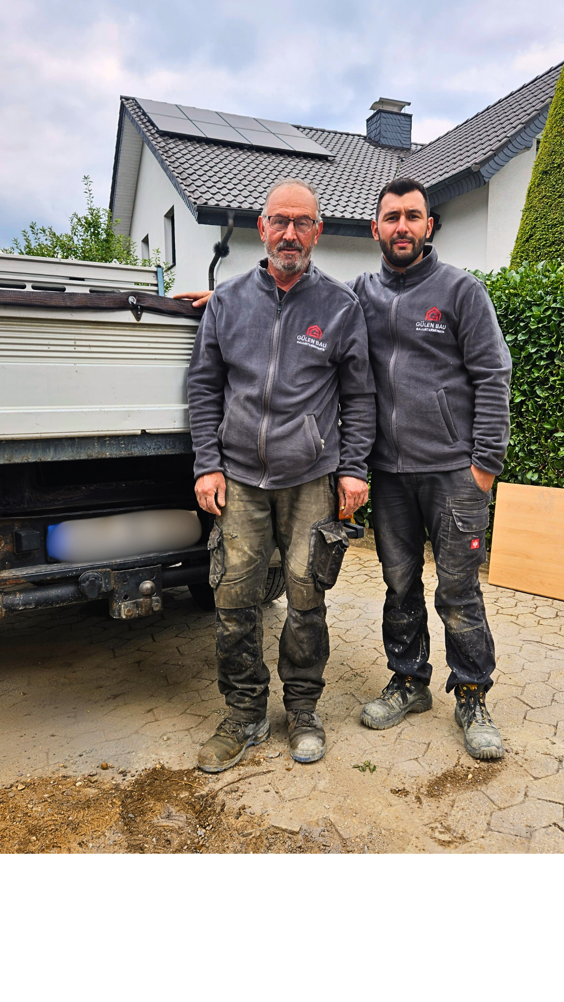 Zwei Männer in Arbeitskleidung vor einem Haus, wahrscheinlich Bauarbeiter oder Handwerker, die neben einem Truck stehen.