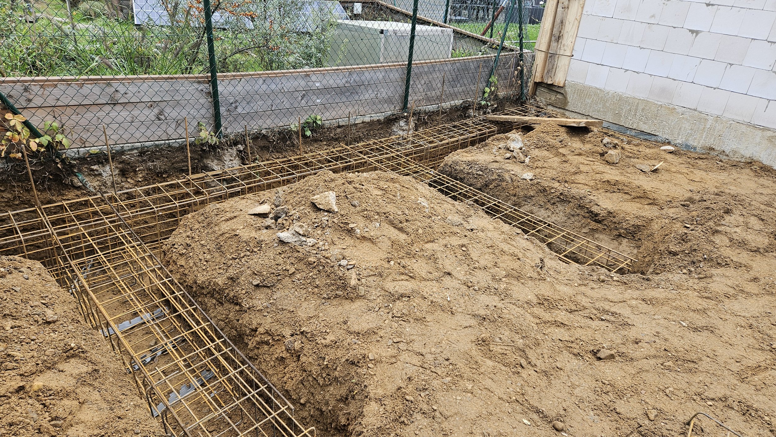 Baustelle mit Eisenstahlgitter und Erdaushebungen vor einem weißen Mauer und Zaun im Garten.