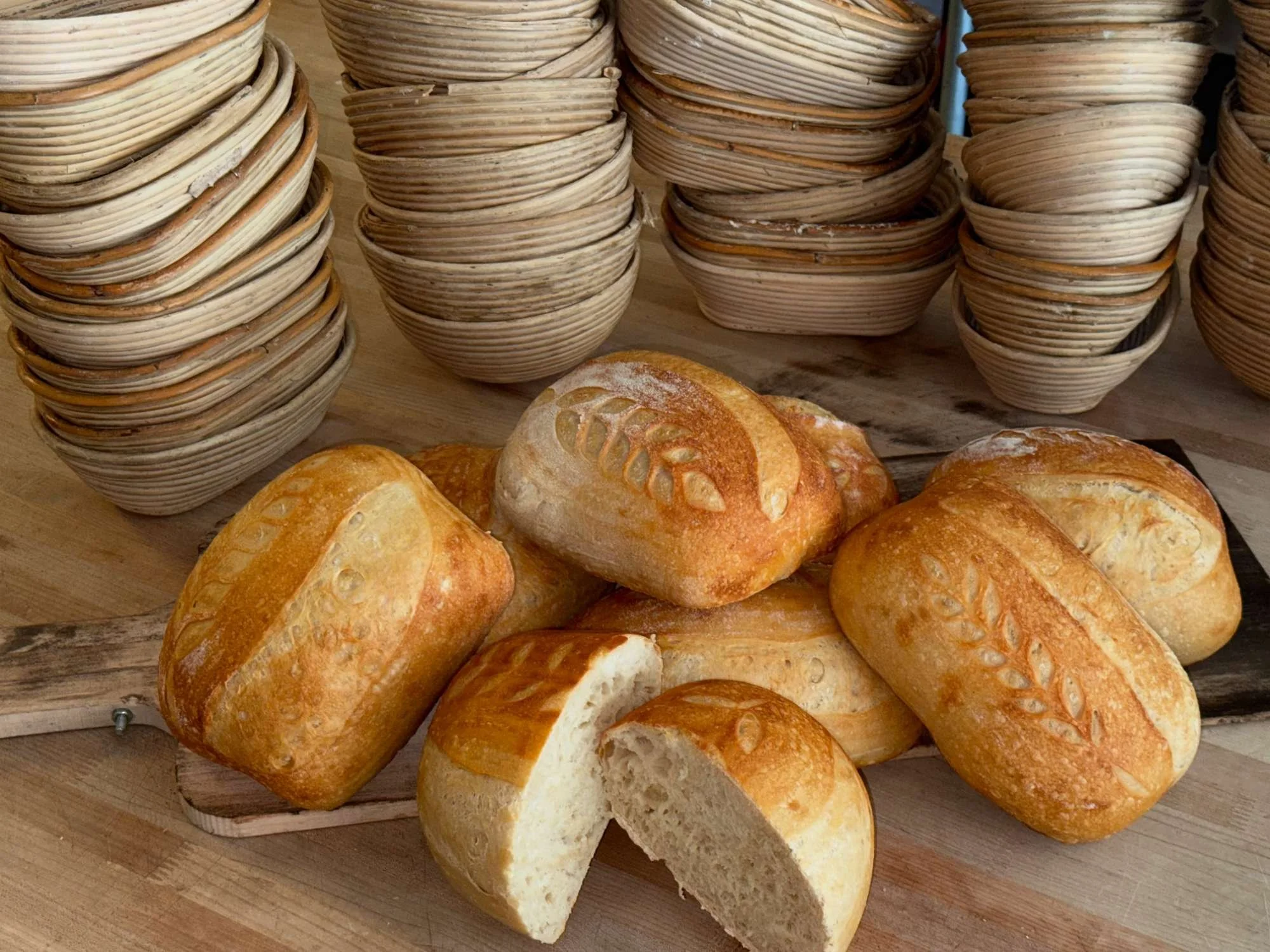 Freshly baked Baxters Artisan Sourdough bread rolls with leaf patterns on top, placed on a wooden board, with stacks of woven baskets in the background.