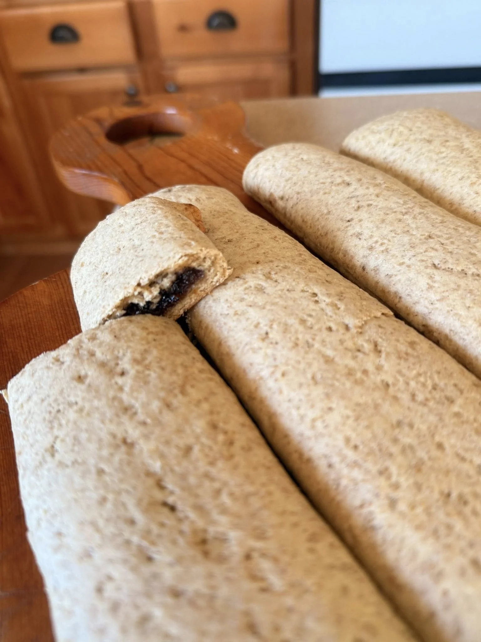 Four rolled Baxters Artisan Sourdough hand pies with one showing a dark filling inside, placed on a wooden cutting board on a kitchen table.