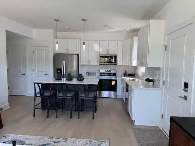 Modern kitchen with white cabinets, stainless steel appliances, a central island with four black chairs, light hardwood floors, and pendant lighting.