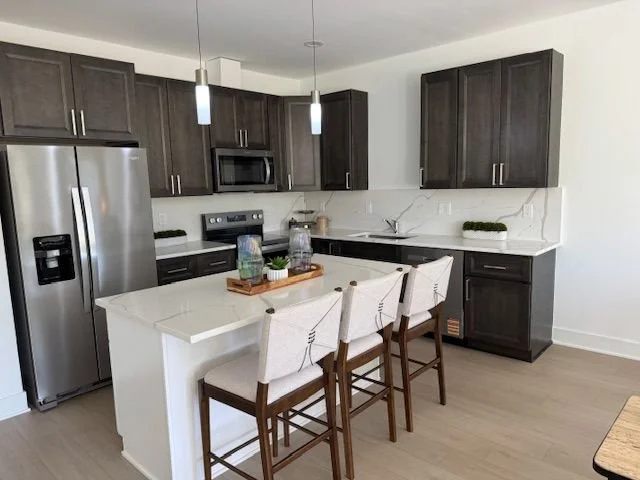 Modern kitchen with dark wood cabinets, stainless steel refrigerator, microwave, and white countertops. Kitchen island with three white barstools, pendant lights above, and decorative items on the island. Light-colored flooring and a white wall backg