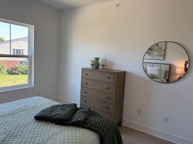 Bedroom with window, dresser with vases, bed with blankets, and round mirror on the wall.