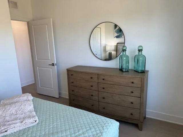 A bedroom with a wooden dresser, a round mirror, two large turquoise glass bottles, a white door slightly open, and part of a bed with a light-colored blanket and mint green quilt.